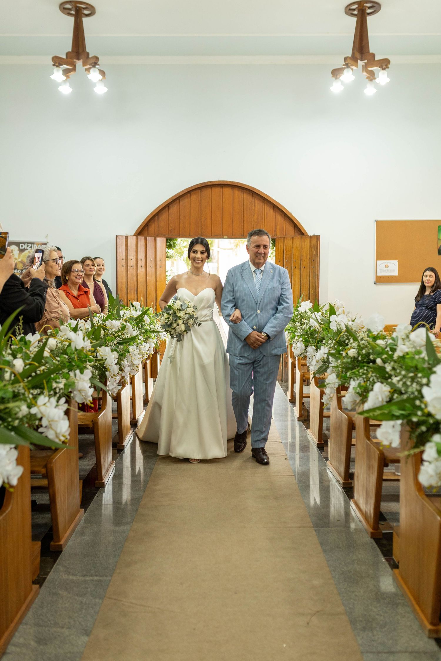 Bride walks down church aisle with escort, flanked by wooden pews adorned with white floral arrangements.