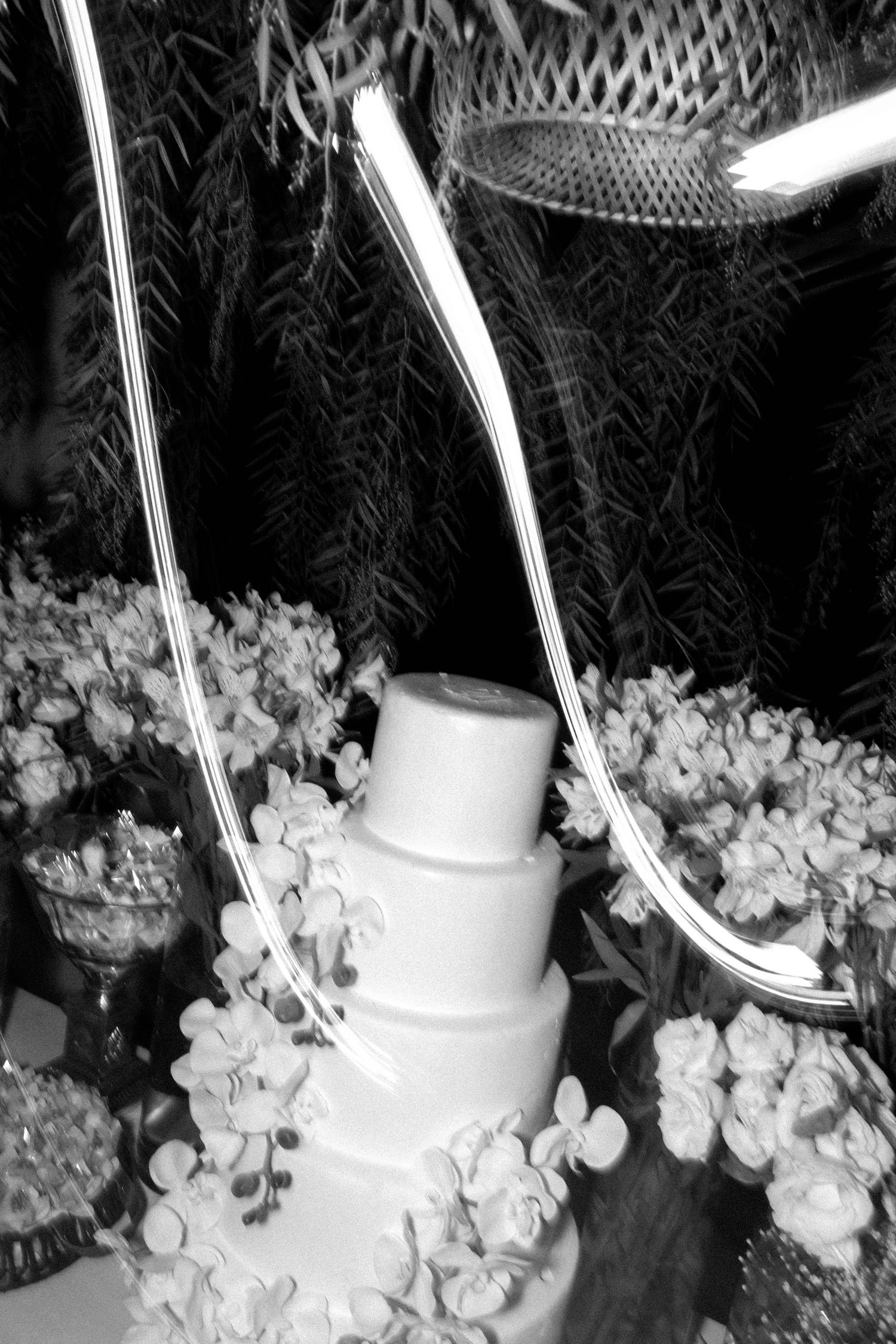 Black and white photo of elegant tiered wedding cake surrounded by flowers with decorative swords crossed above it.