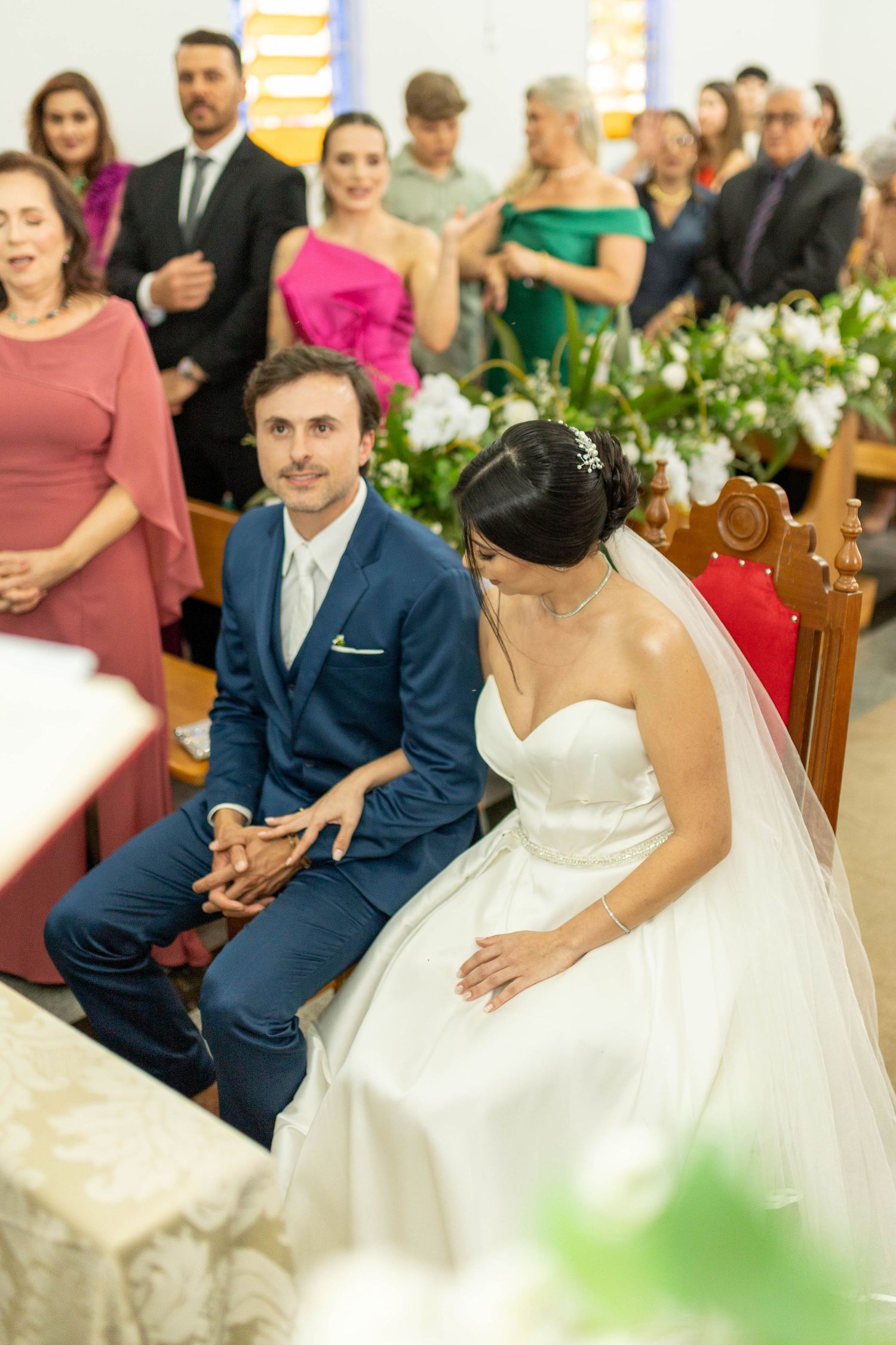 Bride and groom seated at church altar during wedding ceremony, guests standing in background.