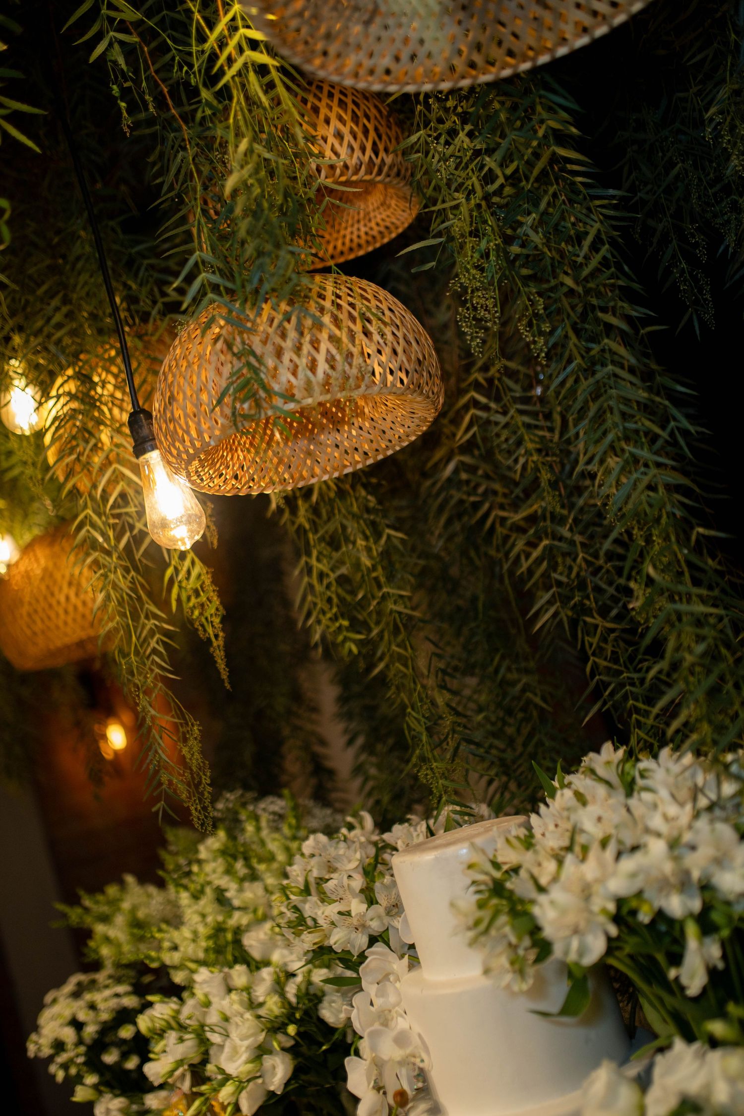 Elegant wedding cake surrounded by white flowers under woven rattan pendant lights and lush green foliage.