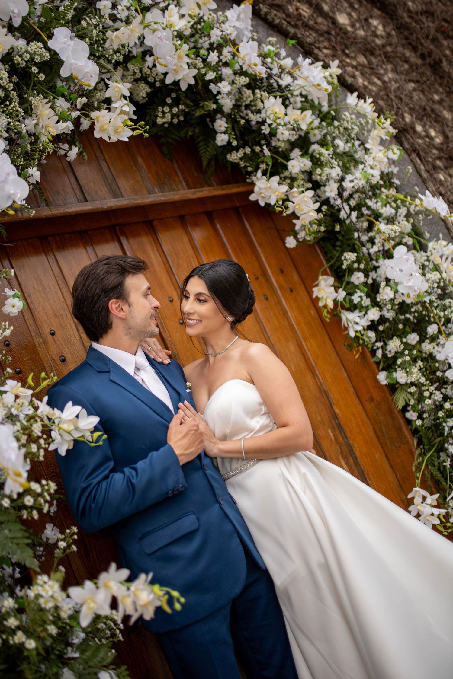 Bride and groom gazing at each other under a white floral arch in front of a wooden door.