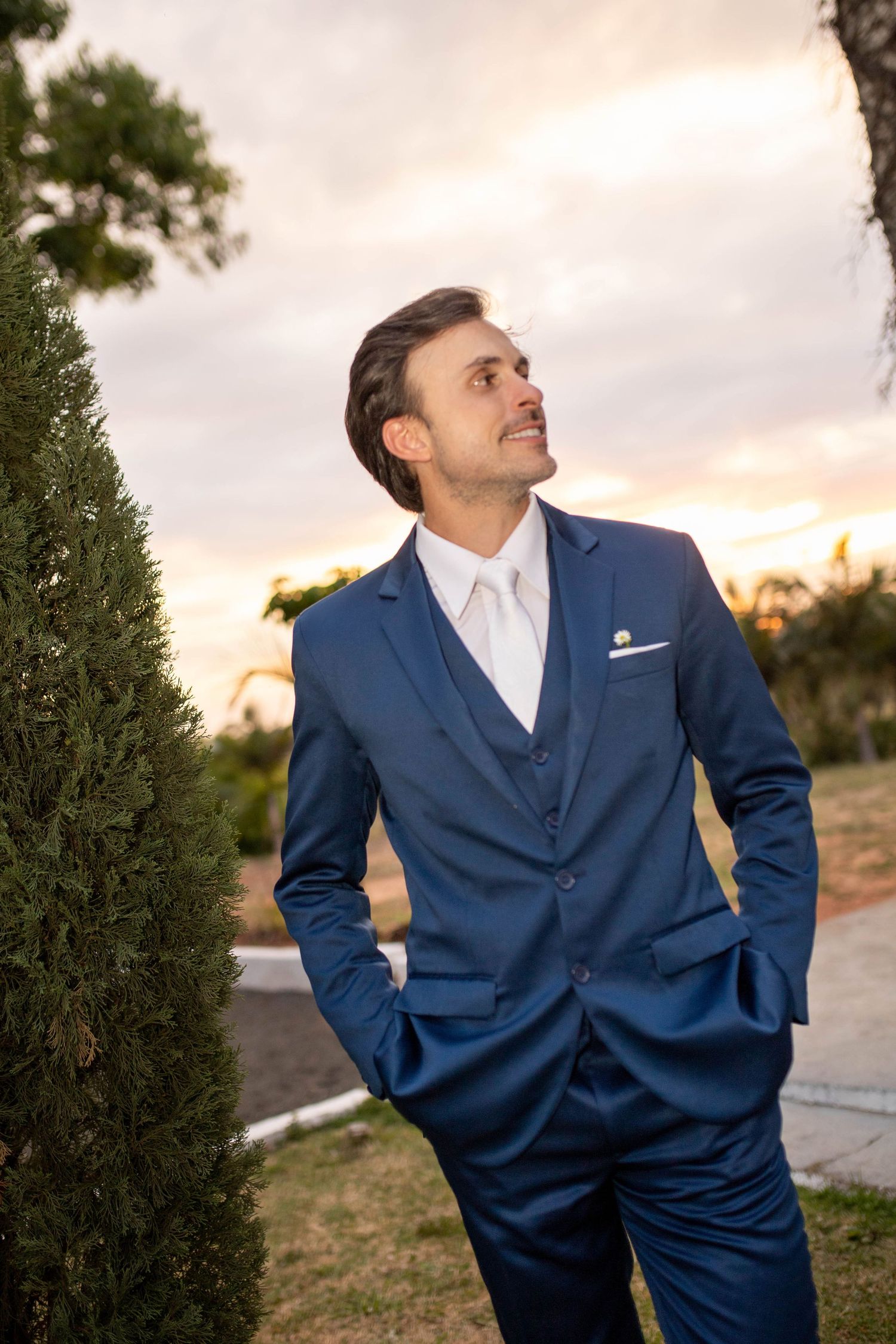 A well-dressed man in a blue three-piece suit with white tie stands outdoors at sunset, hands in pockets.