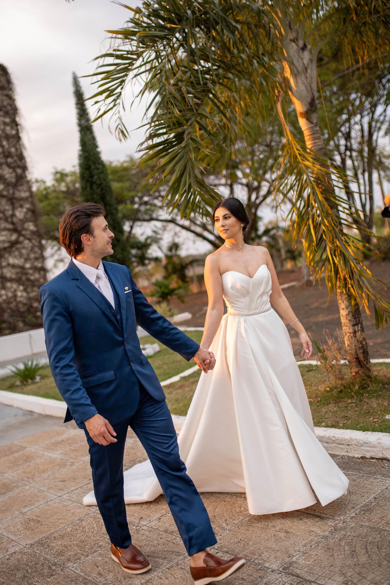 A bride in a white strapless gown and groom in a blue suit hold hands while walking outdoors near palm trees.