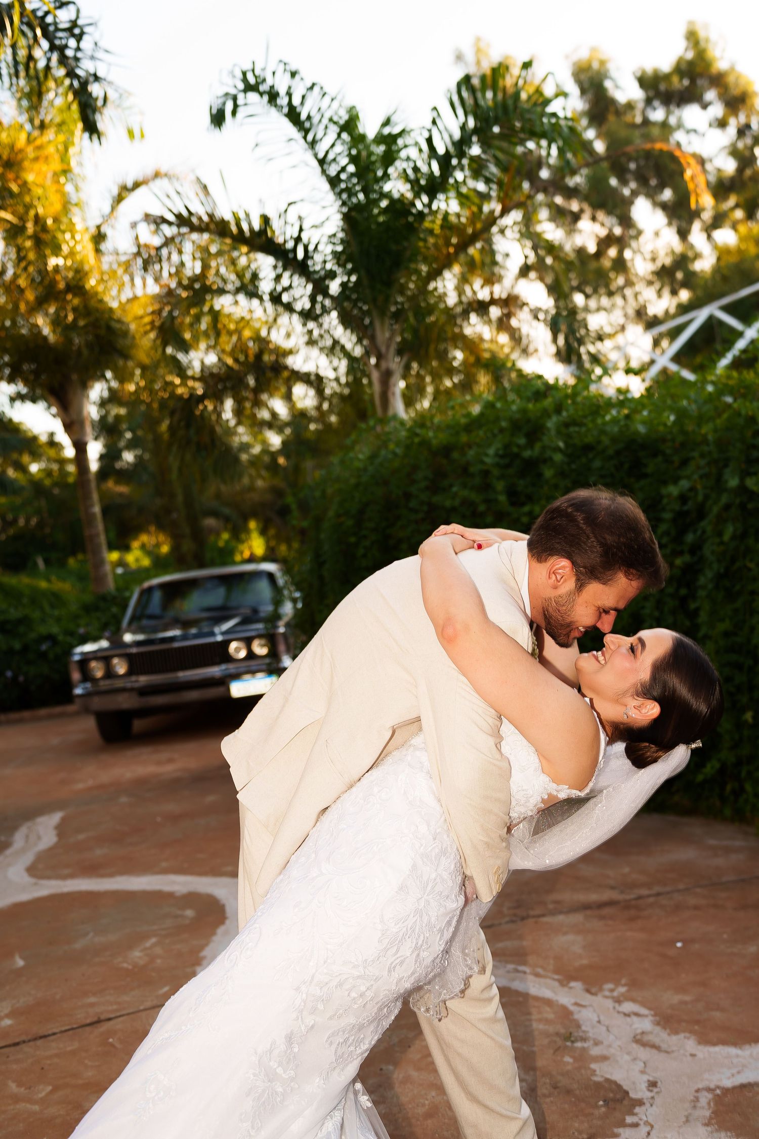 Groom dips bride for a romantic kiss outdoors, surrounded by lush greenery and a classic black car at their wedding.