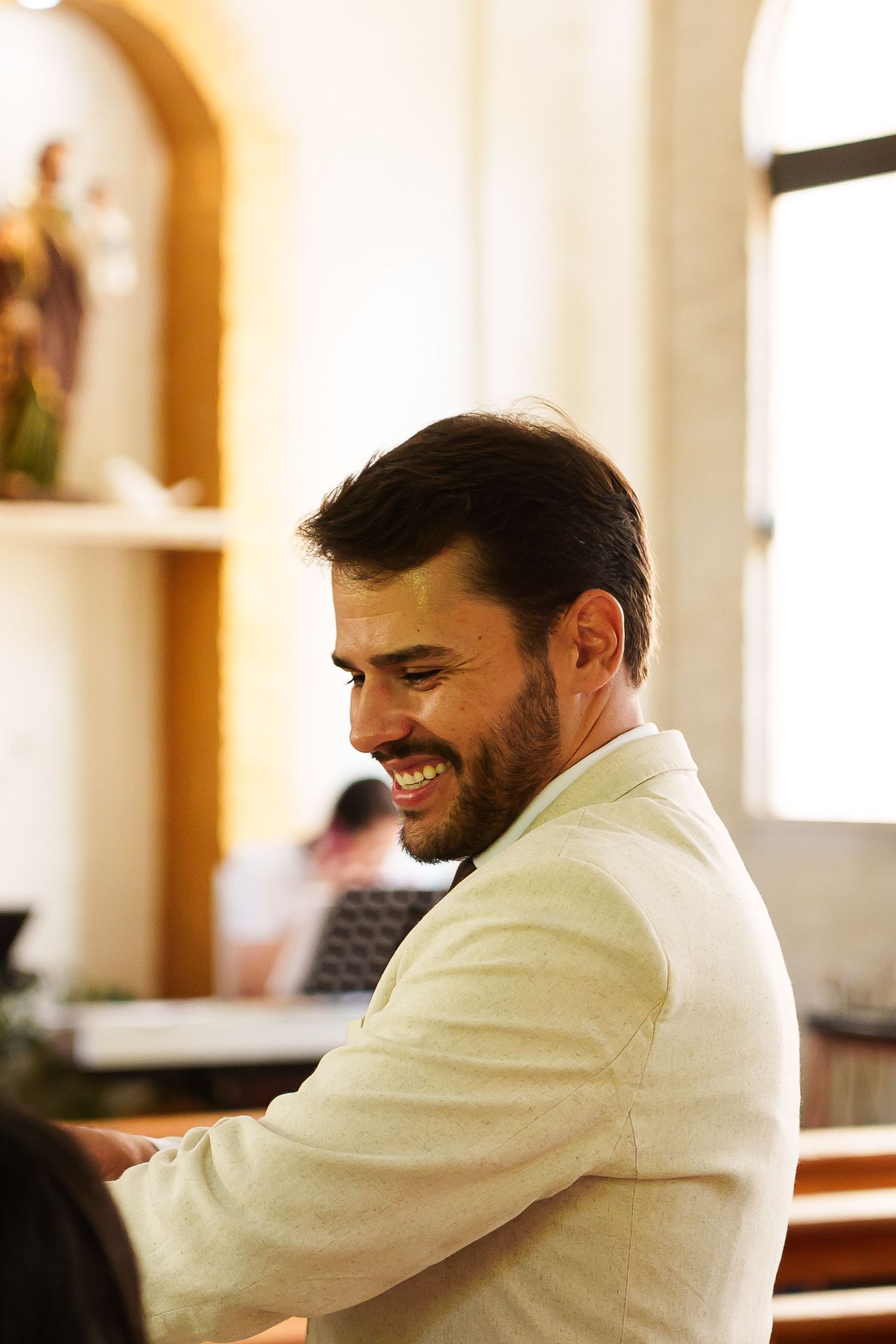 Smiling bearded man in a light suit jacket sitting in a church pew, with religious statues visible in background.