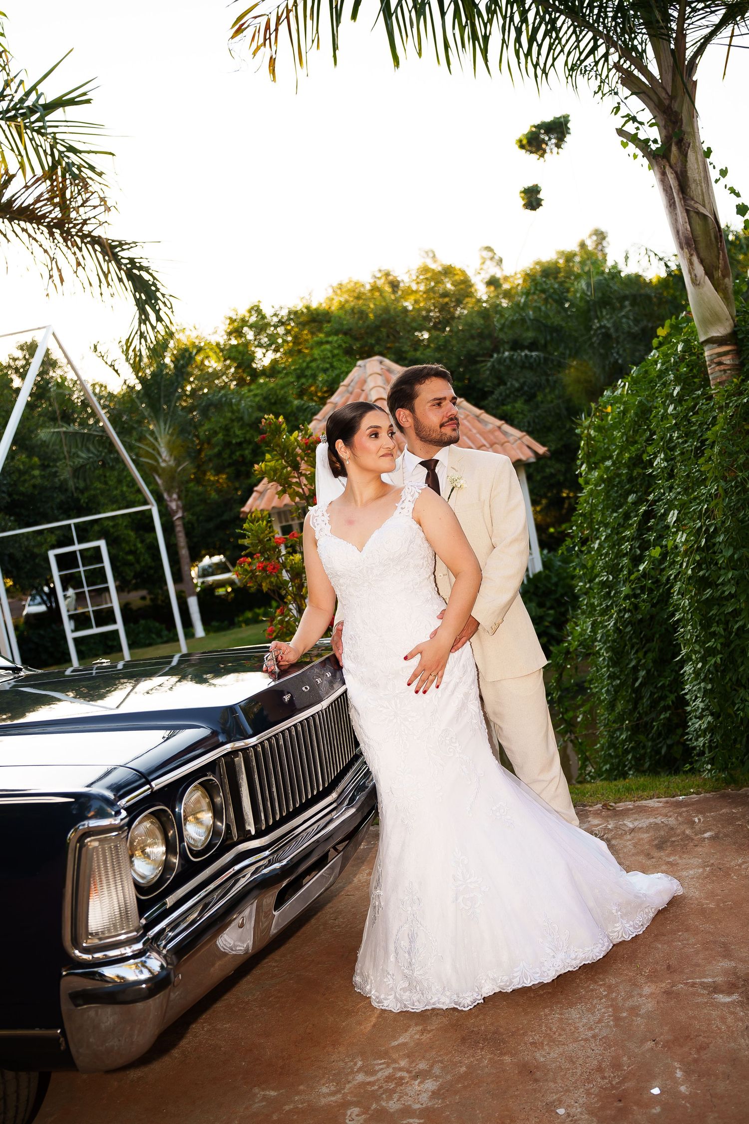 Bride in white lace gown and groom in tan suit pose beside a classic black car surrounded by lush greenery.