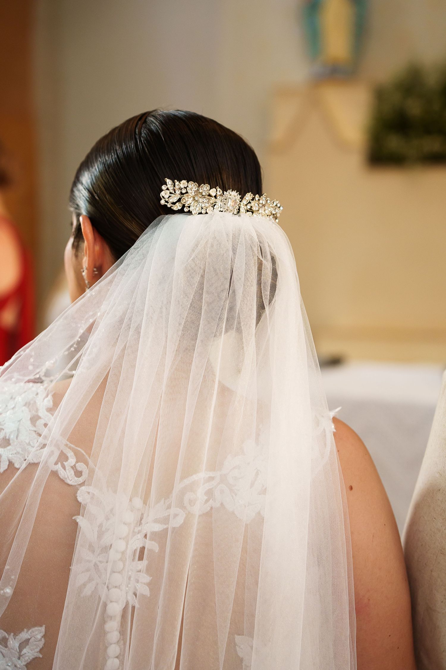 Bride wearing a white lace veil with a crystal hair comb, viewed from behind in a church setting.