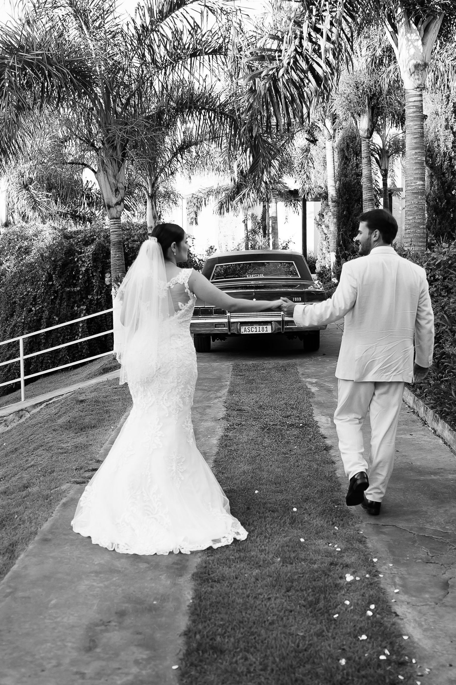 Black and white photo of a bride and groom walking hand in hand toward a classic car surrounded by palm trees.