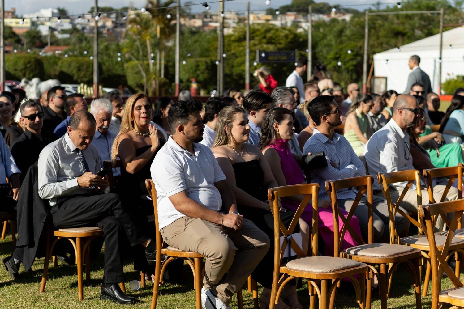 Diverse crowd of people seated in wooden chairs at an outdoor event on a sunny day with green lawn background.
