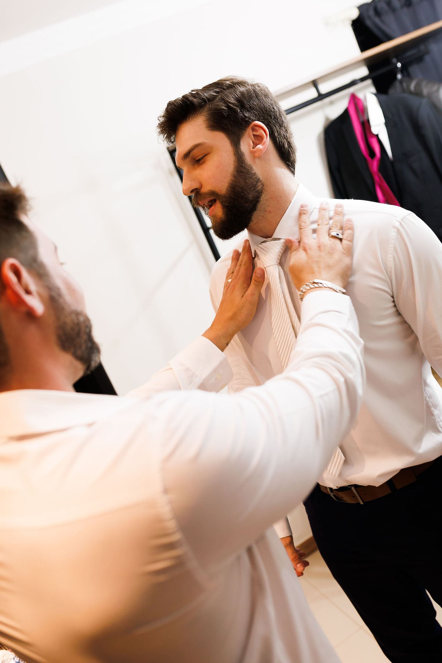 A tailor adjusting a white tie on a bearded man in a suit fitting room.