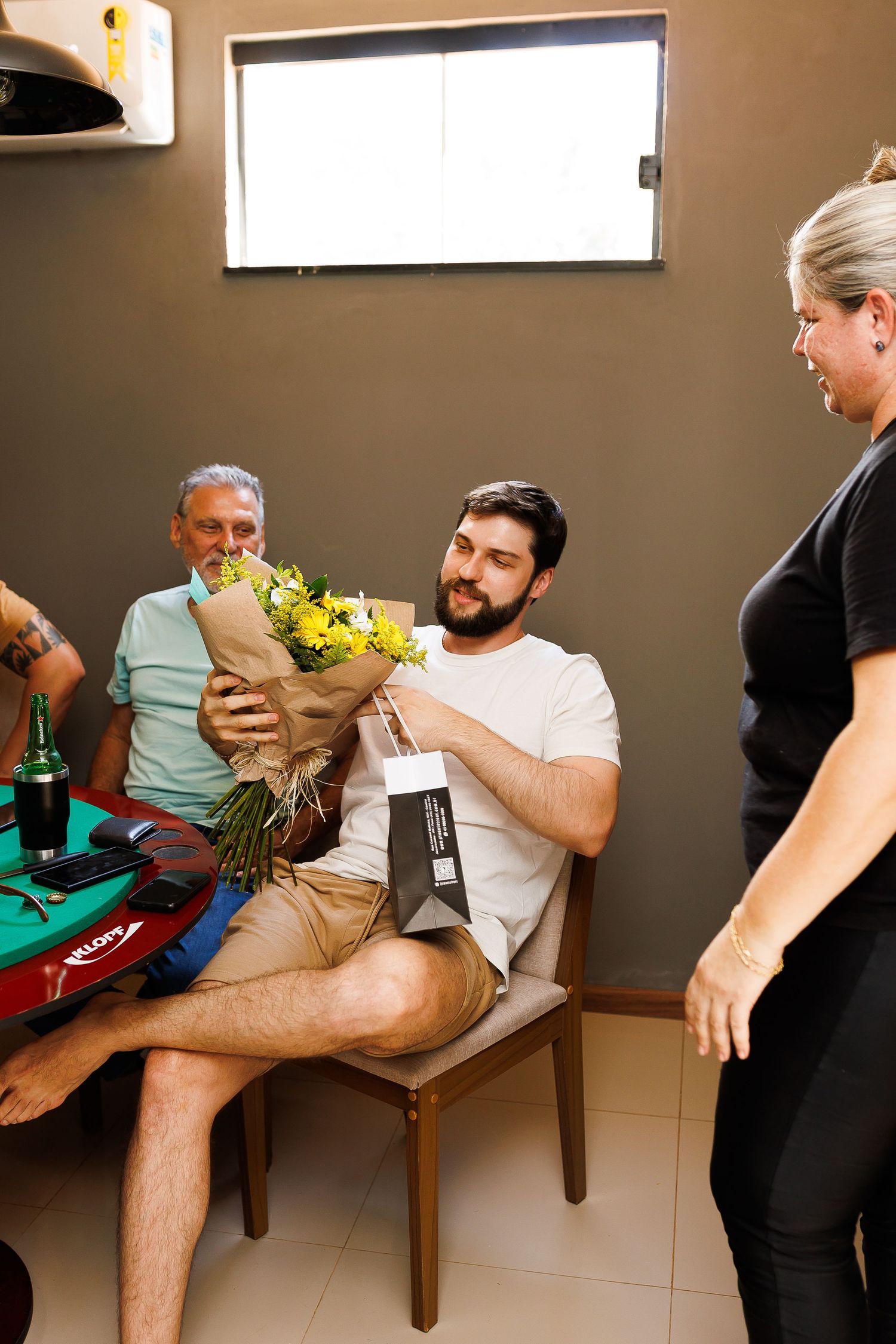 Man with beard receives yellow flower bouquet and gift bag while seated at table with older man nearby.