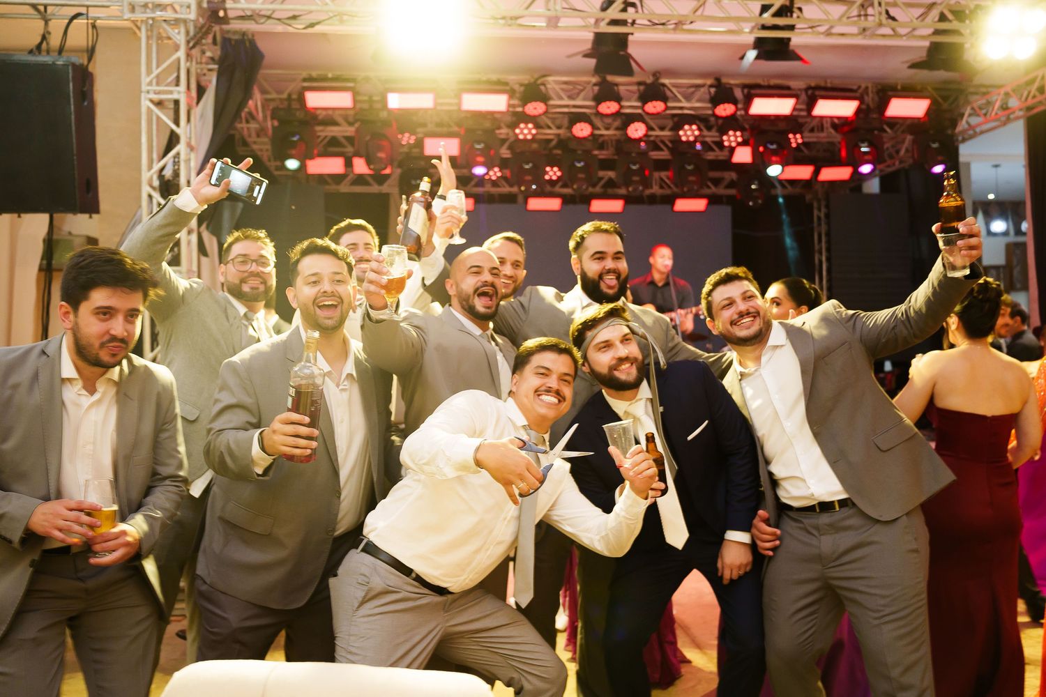 Group of men in suits celebrating at a wedding reception, laughing and taking selfies under stage lights.