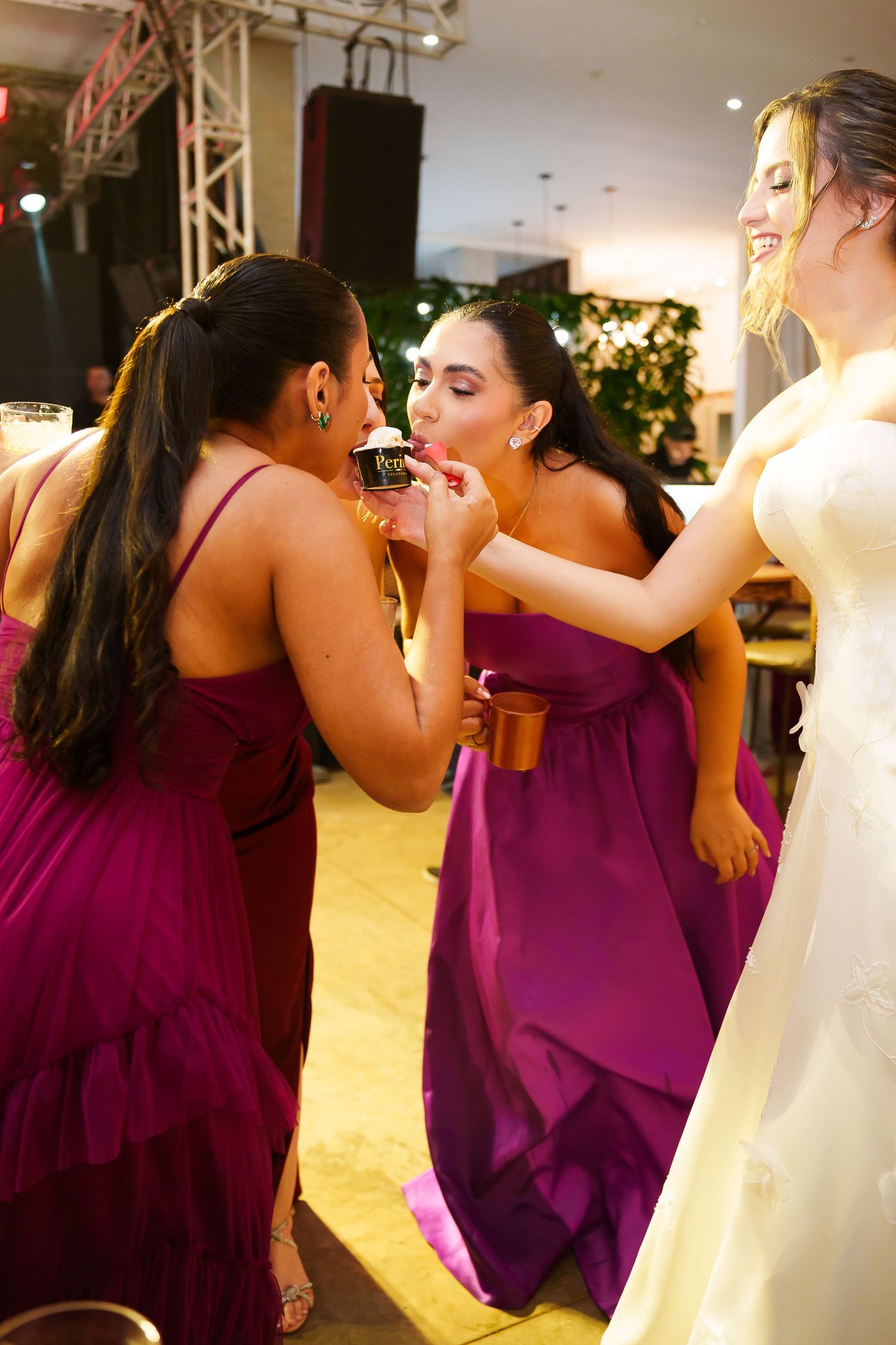 Bridesmaids in purple dresses sharing drinks at a wedding reception while the bride in white looks on.