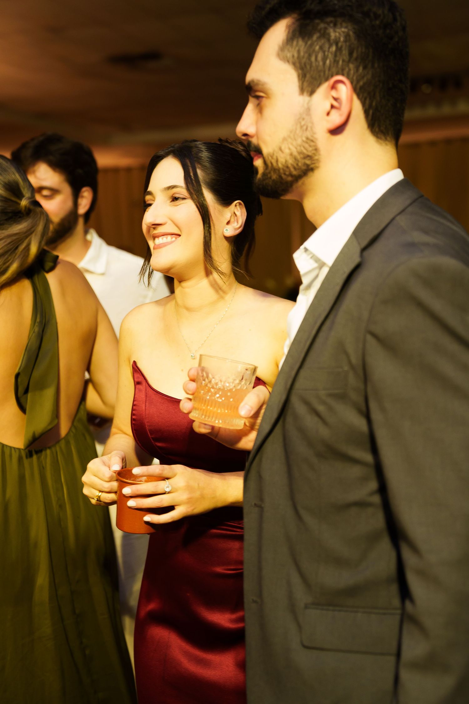 Elegant couple at a party, woman in red dress holding a cocktail glass, smiling at bearded man in gray suit.