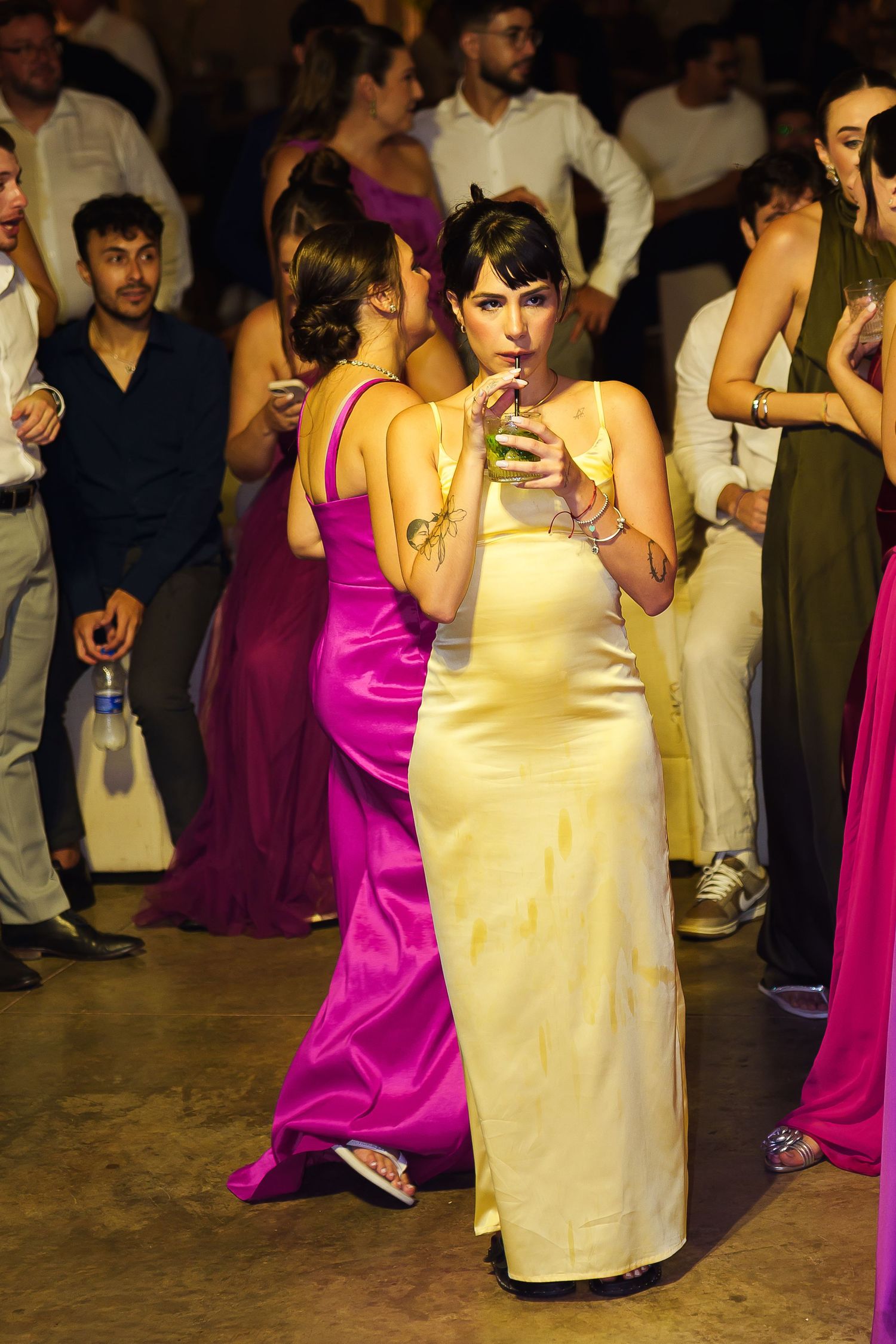 Woman in yellow satin gown sipping cocktail on dance floor at elegant party with guests in formal attire.