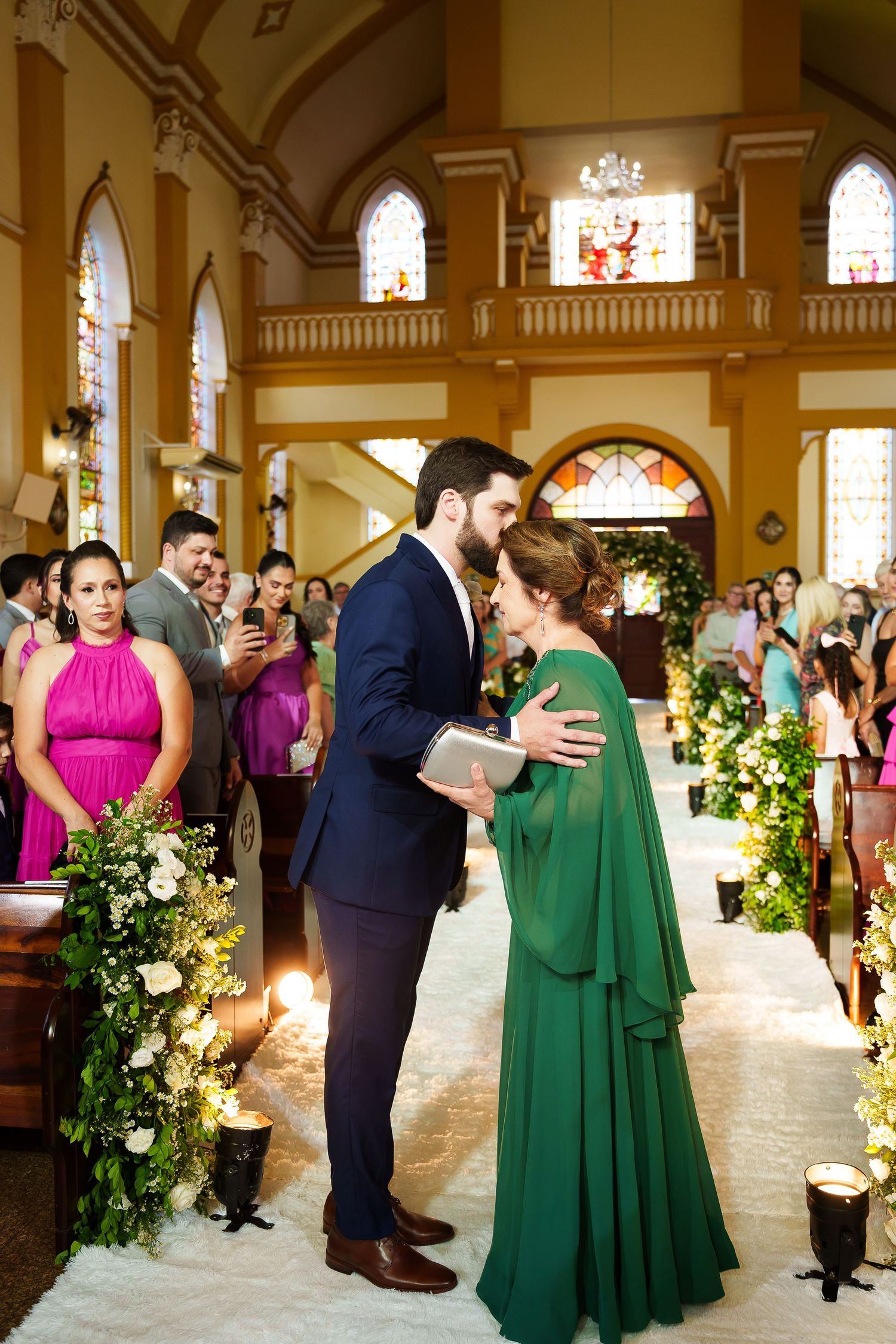 A groom in a navy suit greets a woman in an elegant green cape dress at a floral-adorned church wedding aisle.
