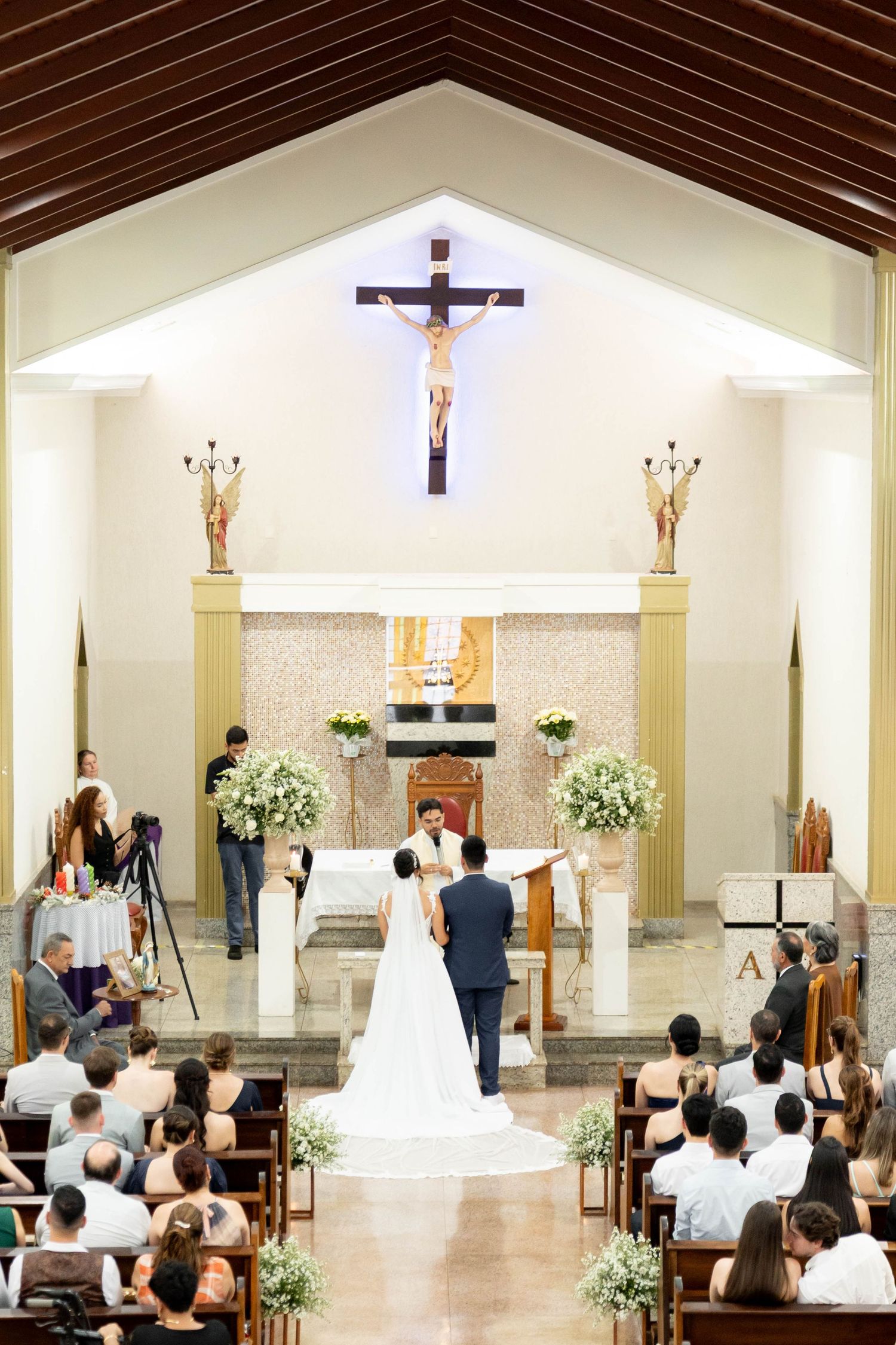 Bride and groom exchange vows at Catholic church altar with crucifix, flanked by floral arrangements and wedding guests.