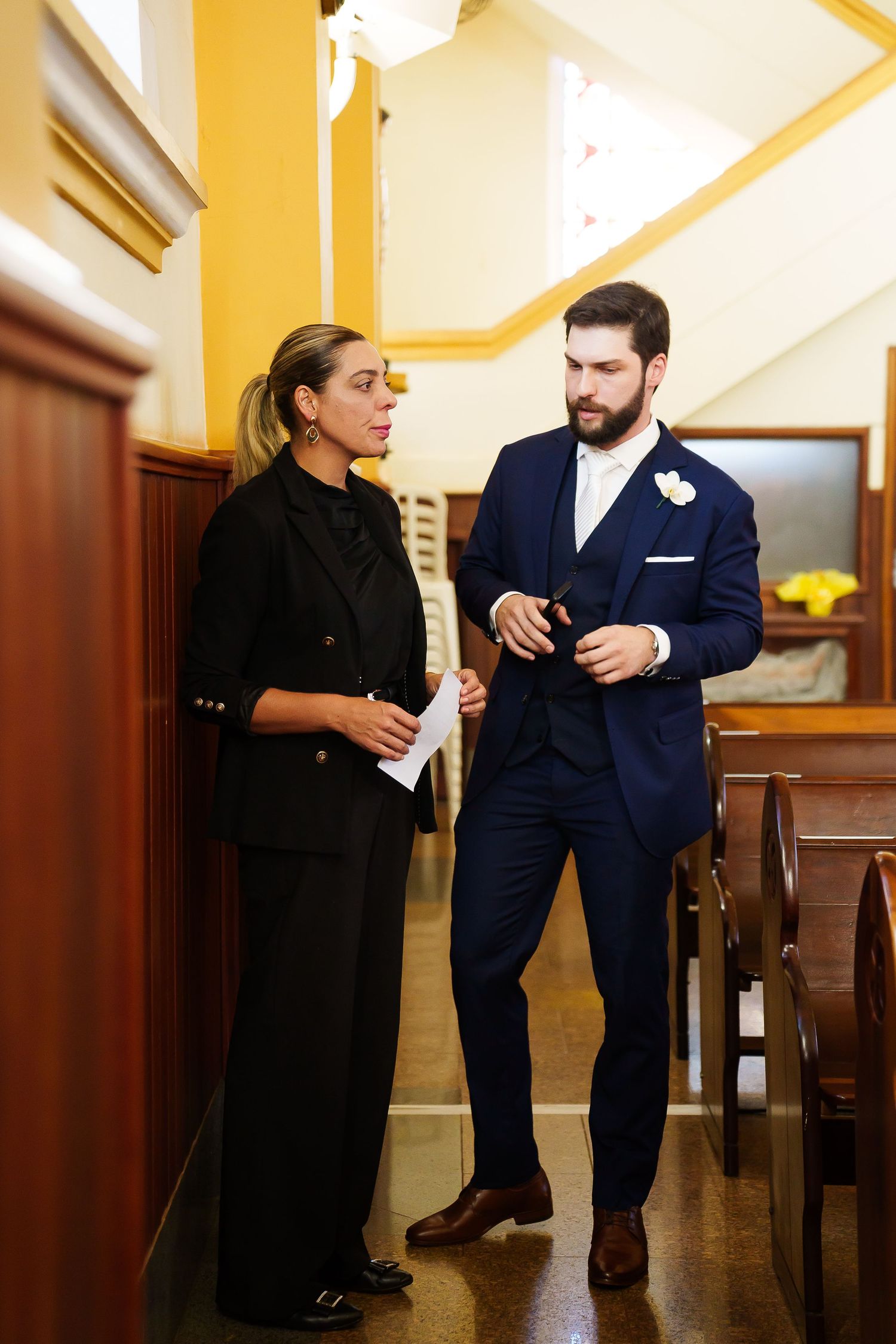 Groom in navy suit with boutonniere consulting with wedding coordinator in black outfit inside a church.