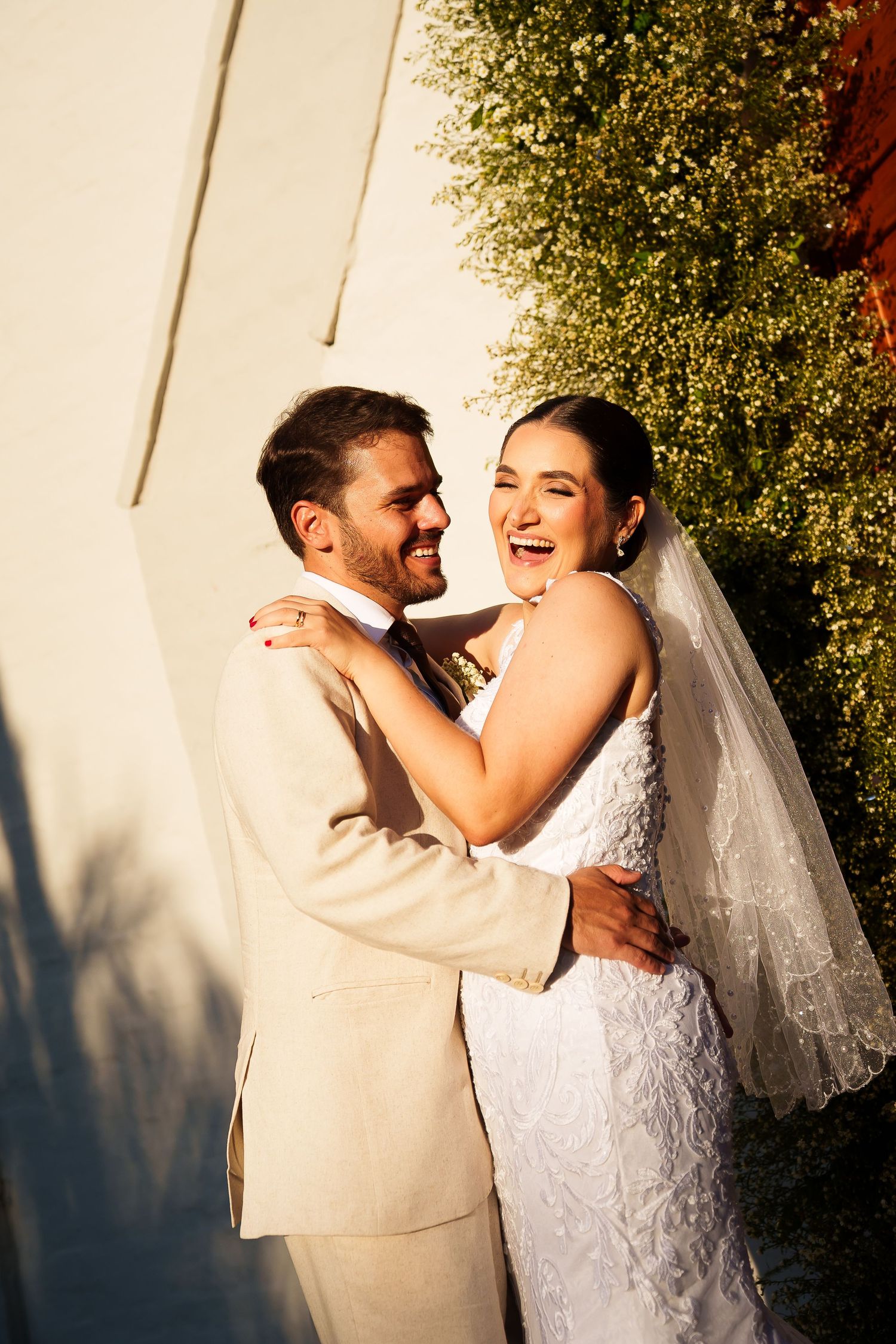 Joyful bride in white lace gown and veil laughing with groom in beige suit outdoors in golden sunlight.
