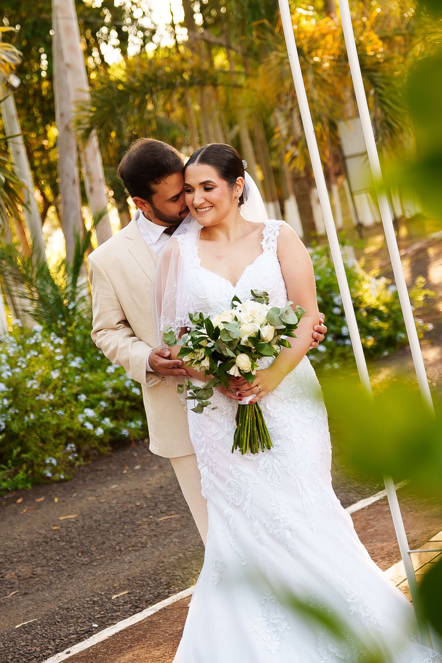Bride and groom embracing in garden, bride holding white rose bouquet in lace wedding dress.