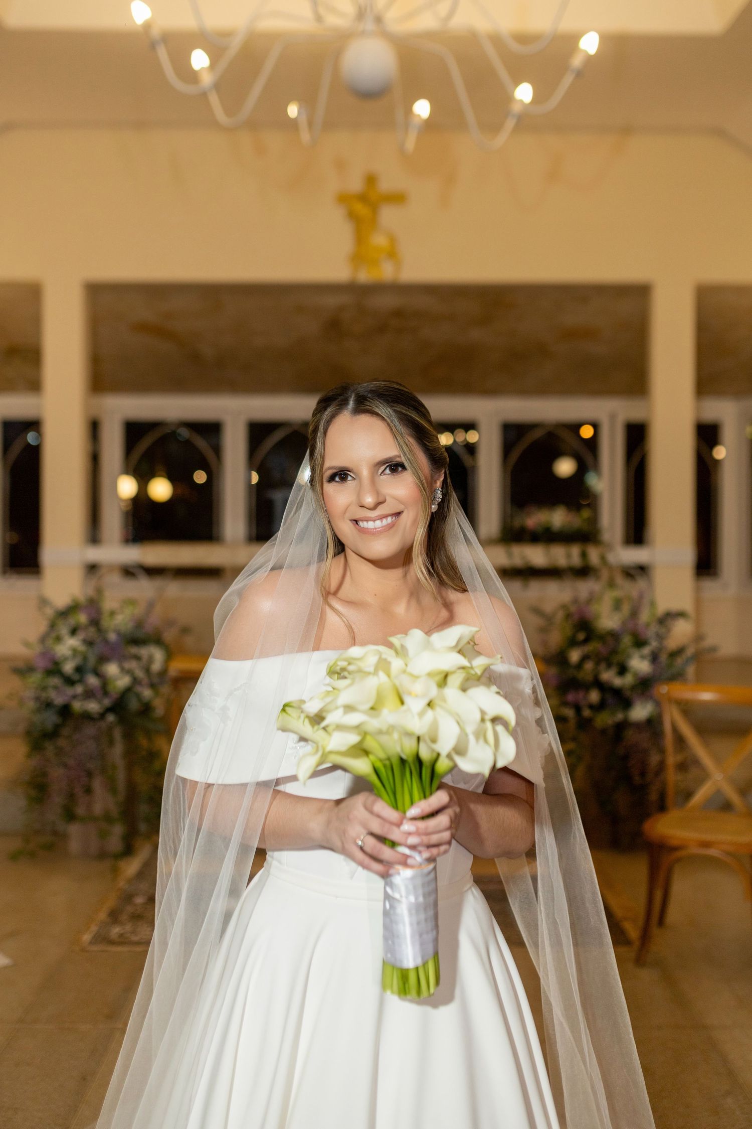 Smiling bride in off-shoulder white gown holds calla lily bouquet with veil, standing in elegant church interior.