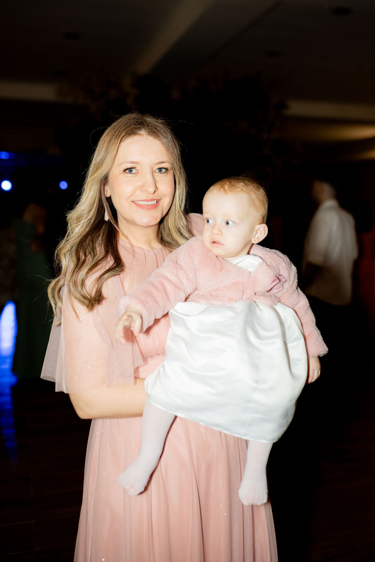 Woman in pink dress holding a baby in white outfit at an indoor event.