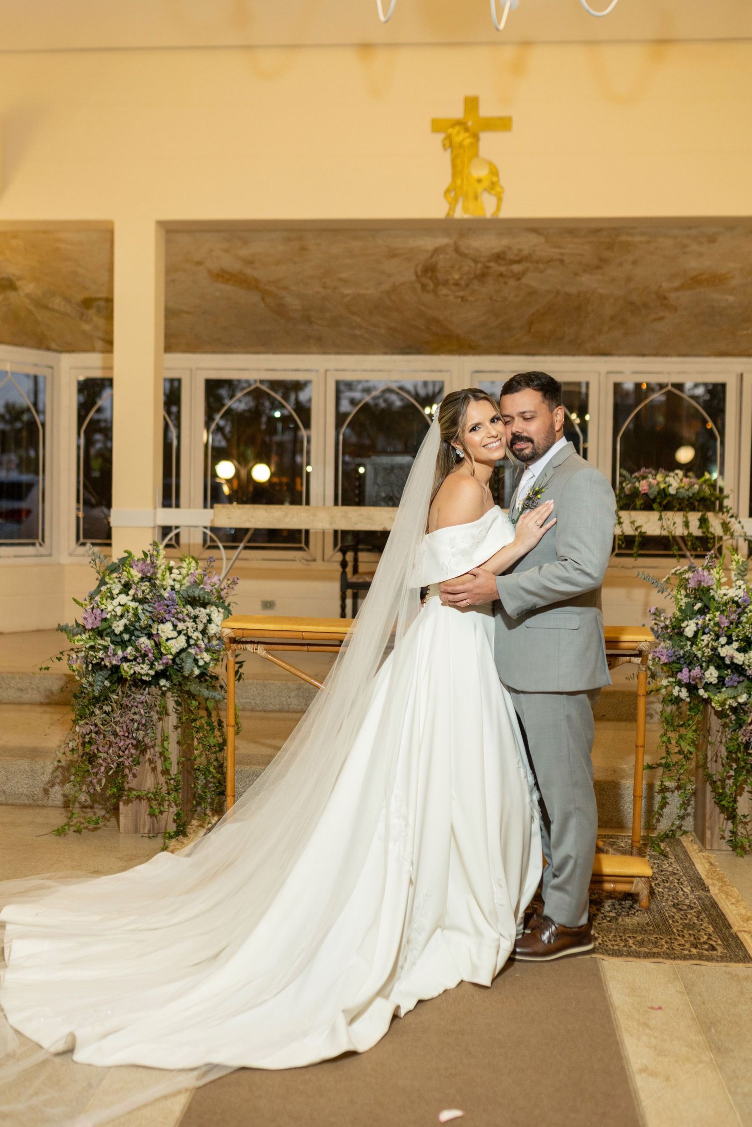 Bride in white gown with long veil and groom in gray suit embrace at church altar with floral arrangements.