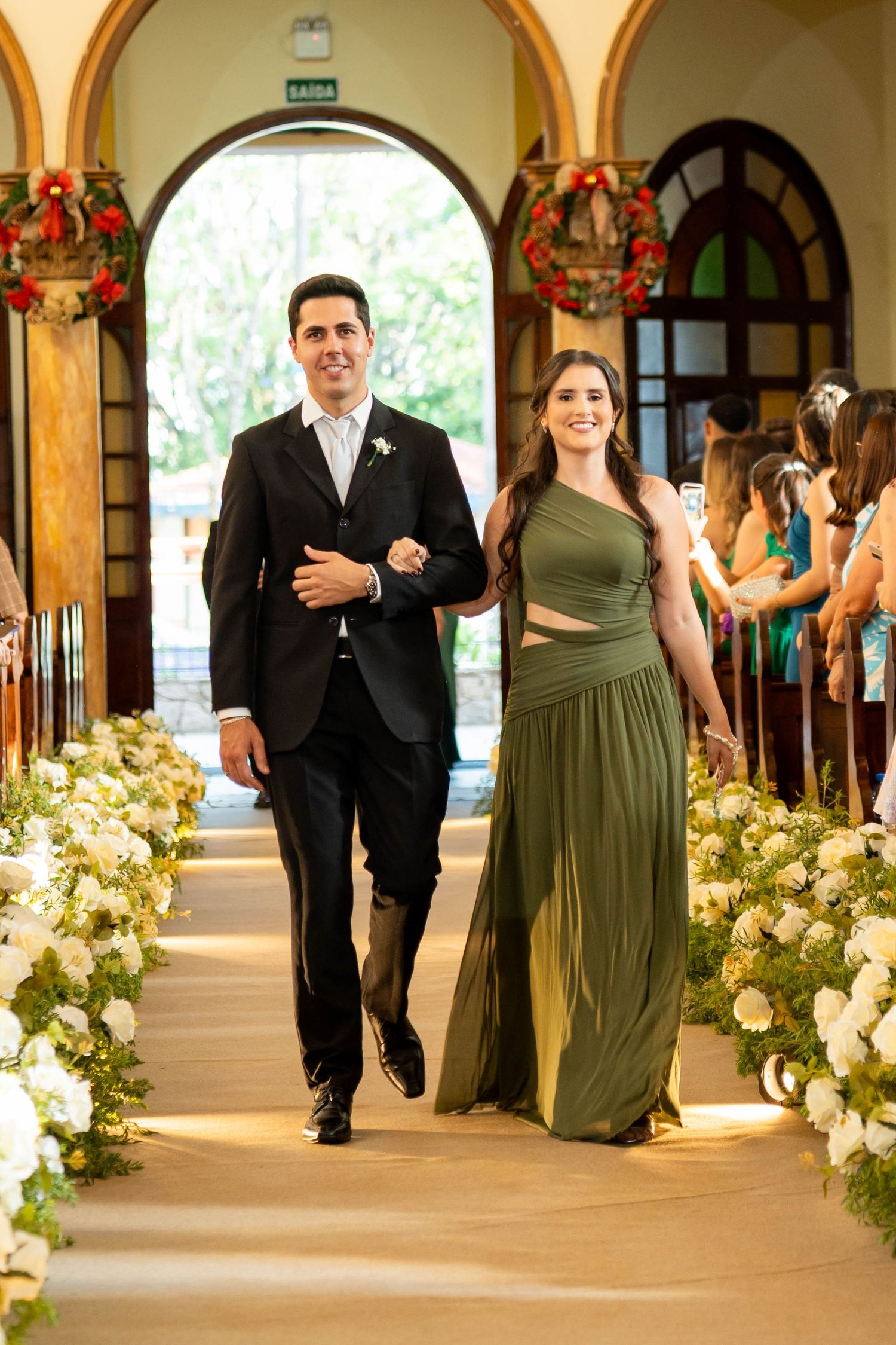 A man in a black suit and woman in an olive green gown walk down a flower-lined church aisle at a wedding ceremony.