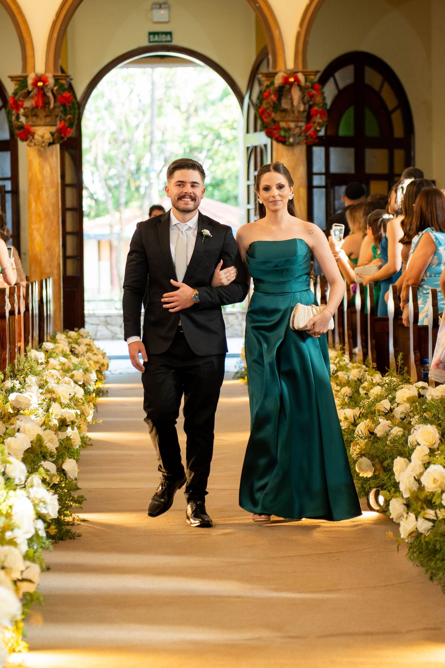 Couple walking down flower-lined church aisle, man in black suit and woman in elegant emerald green gown at wedding ceremony.