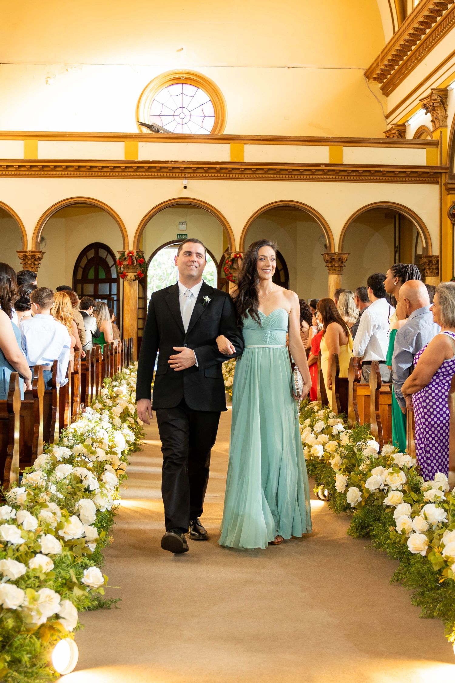 A man in a black tuxedo and woman in a mint green gown walk down a floral-lined church aisle at a wedding.