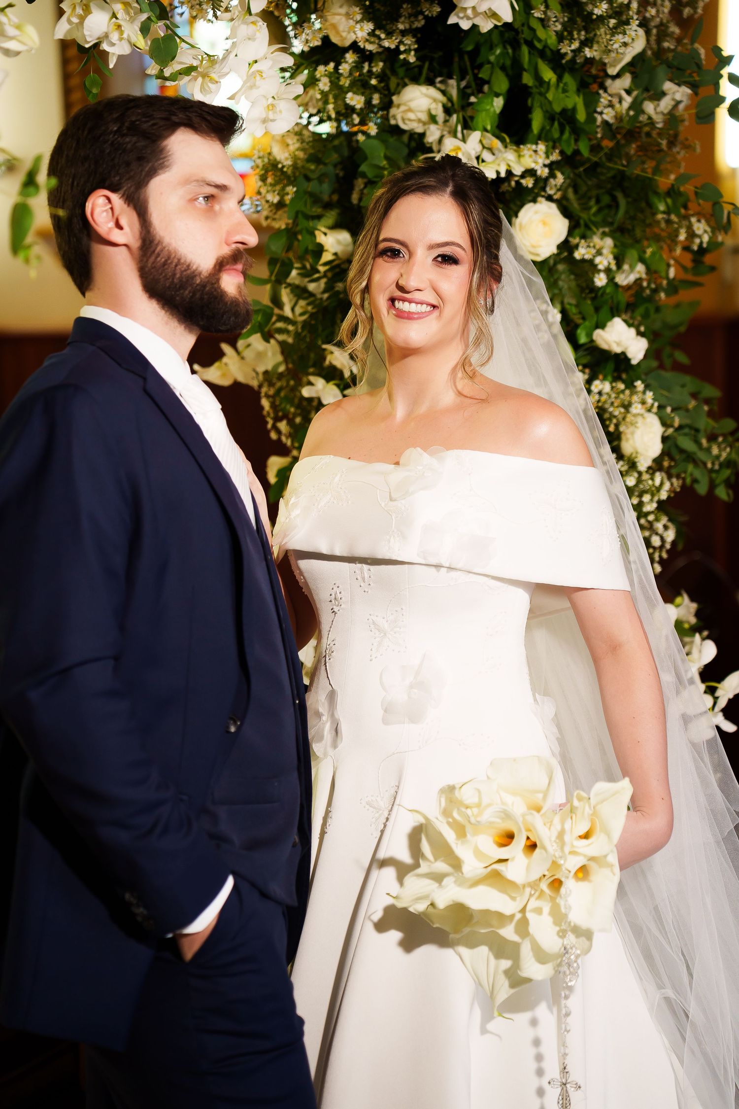 Bride in white off-shoulder gown holding calla lily bouquet stands with bearded groom in navy suit at floral altar.