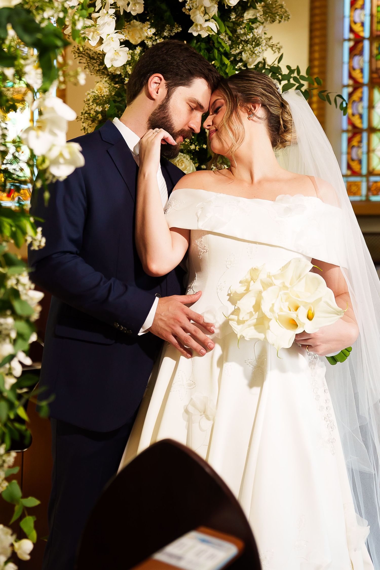 Bride and groom share an intimate moment at church altar, bride in off-shoulder gown holding white floral bouquet.
