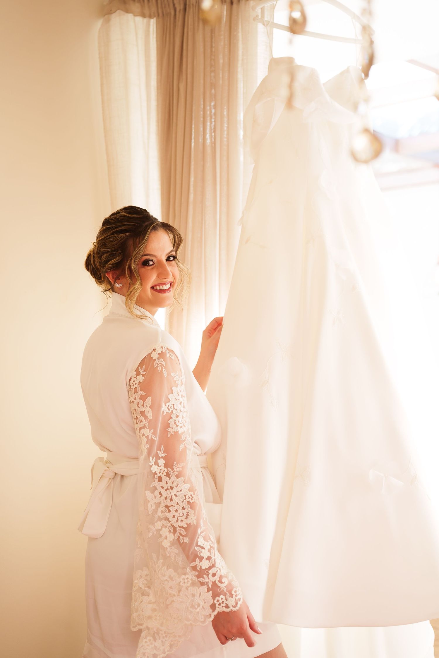 Smiling bride in lace robe admiring her white wedding dress hanging by a sunlit window.
