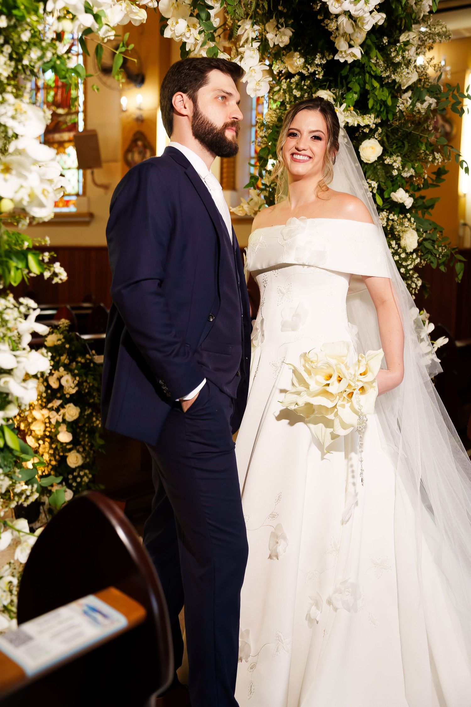 Bride in off-shoulder white gown holding bouquet stands with bearded groom in navy suit at church wedding ceremony.