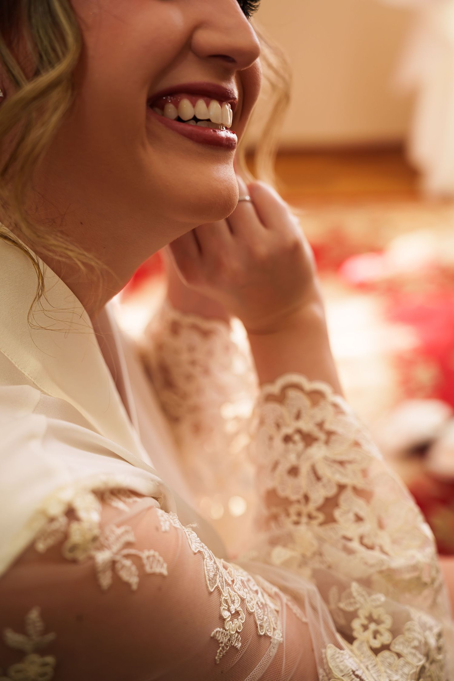 Smiling bride in lace wedding robe touching her face, showcasing elegant floral lace sleeves and a joyful expression.