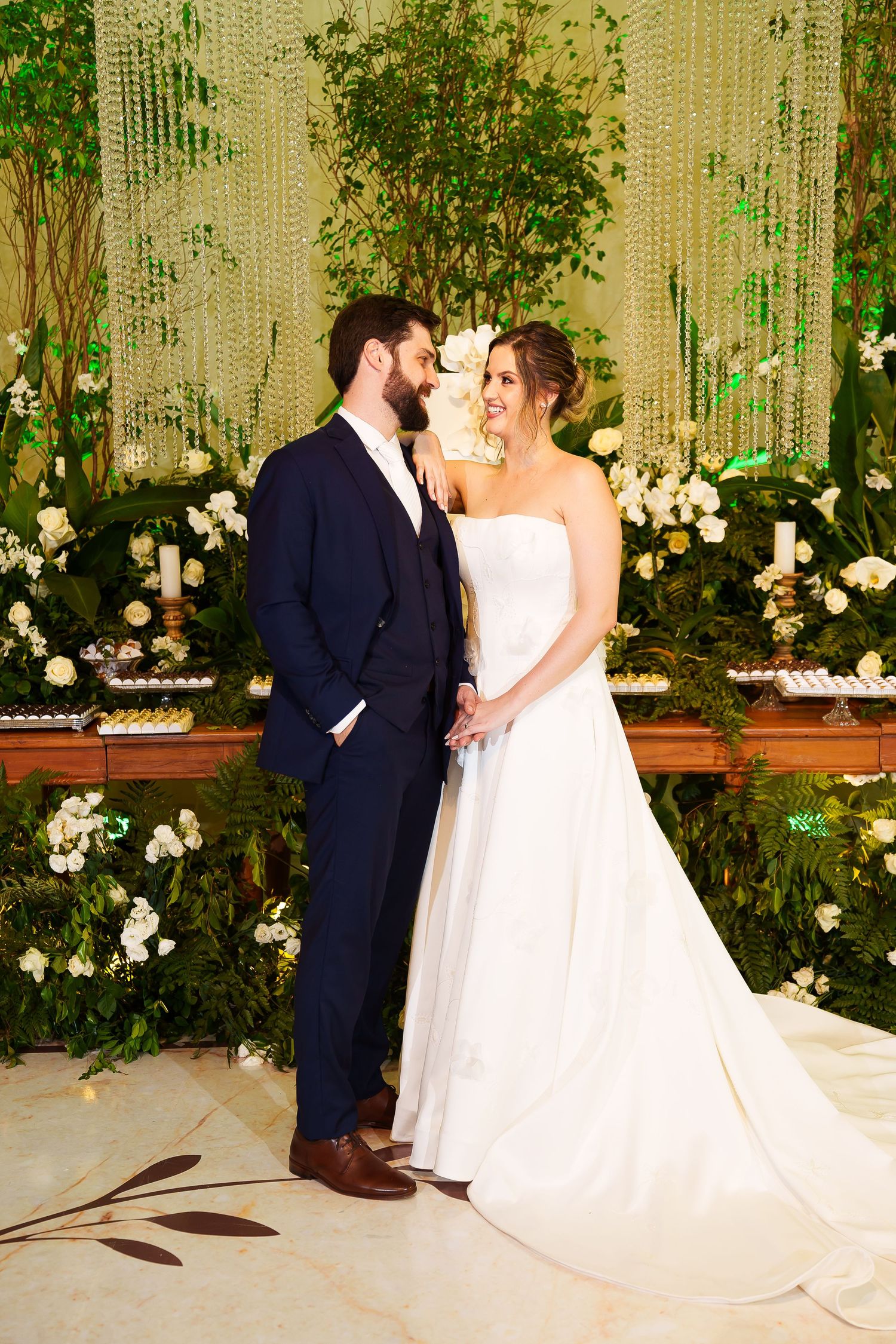 Bride in strapless white gown and groom in navy suit share a romantic moment at floral wedding reception.