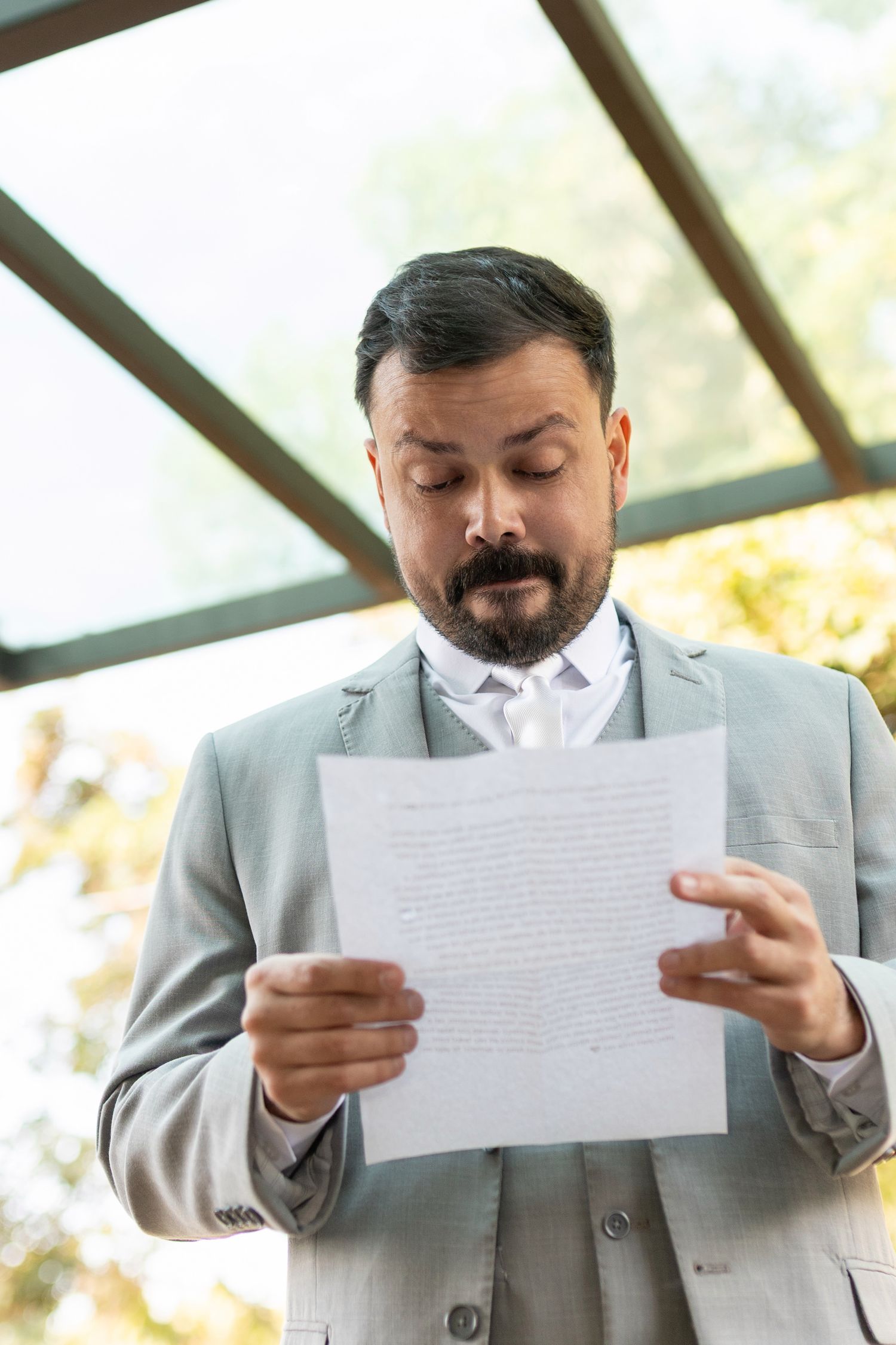 Businessman in gray suit reading a document with a serious expression, standing indoors near large glass windows.