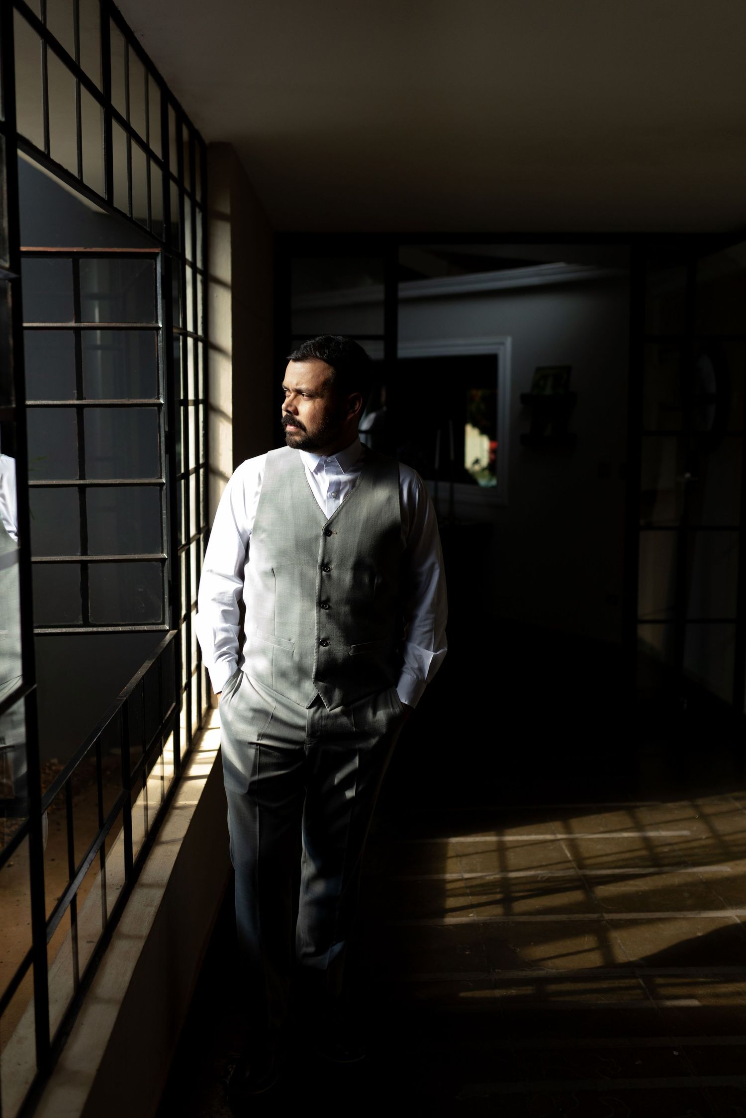 Man in gray vest and white shirt stands in dramatic light and shadow near window in dark hallway.