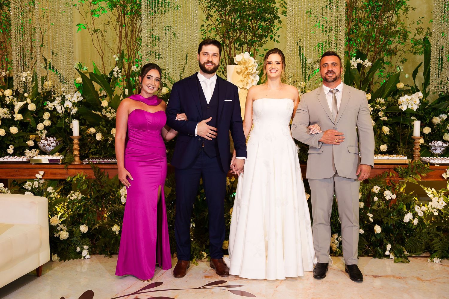 Wedding party of four posing elegantly before lush floral backdrop, bride in white gown with groom in navy suit.