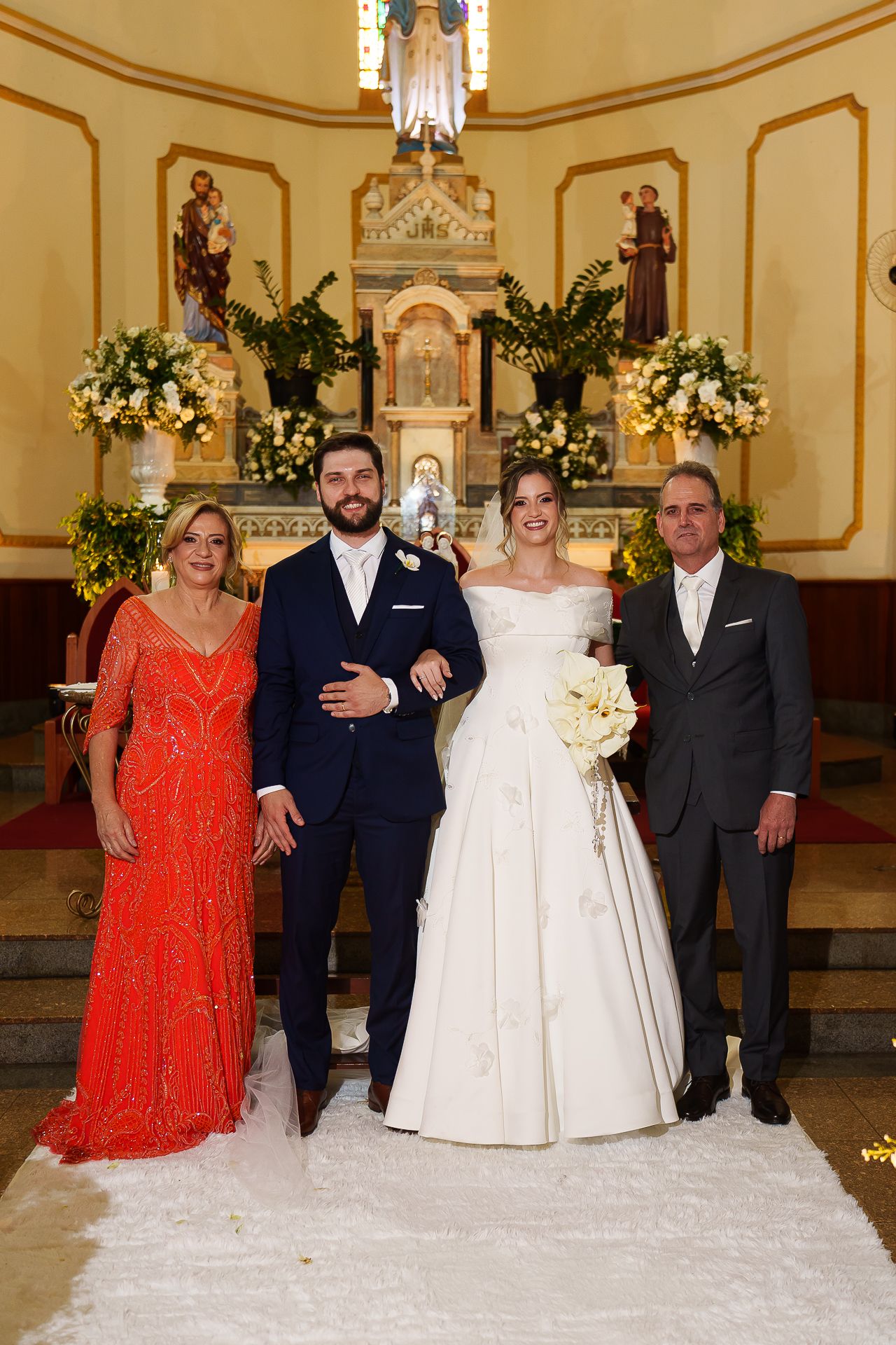 Bride and groom pose with family at church altar, bride in white gown holding bouquet, mother in red dress.