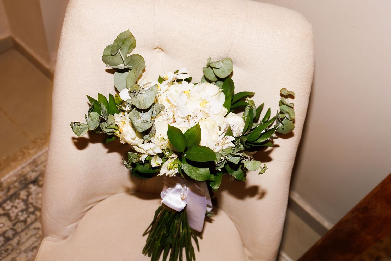 White peony and eucalyptus wedding bouquet with lavender ribbon resting on a cream upholstered chair.