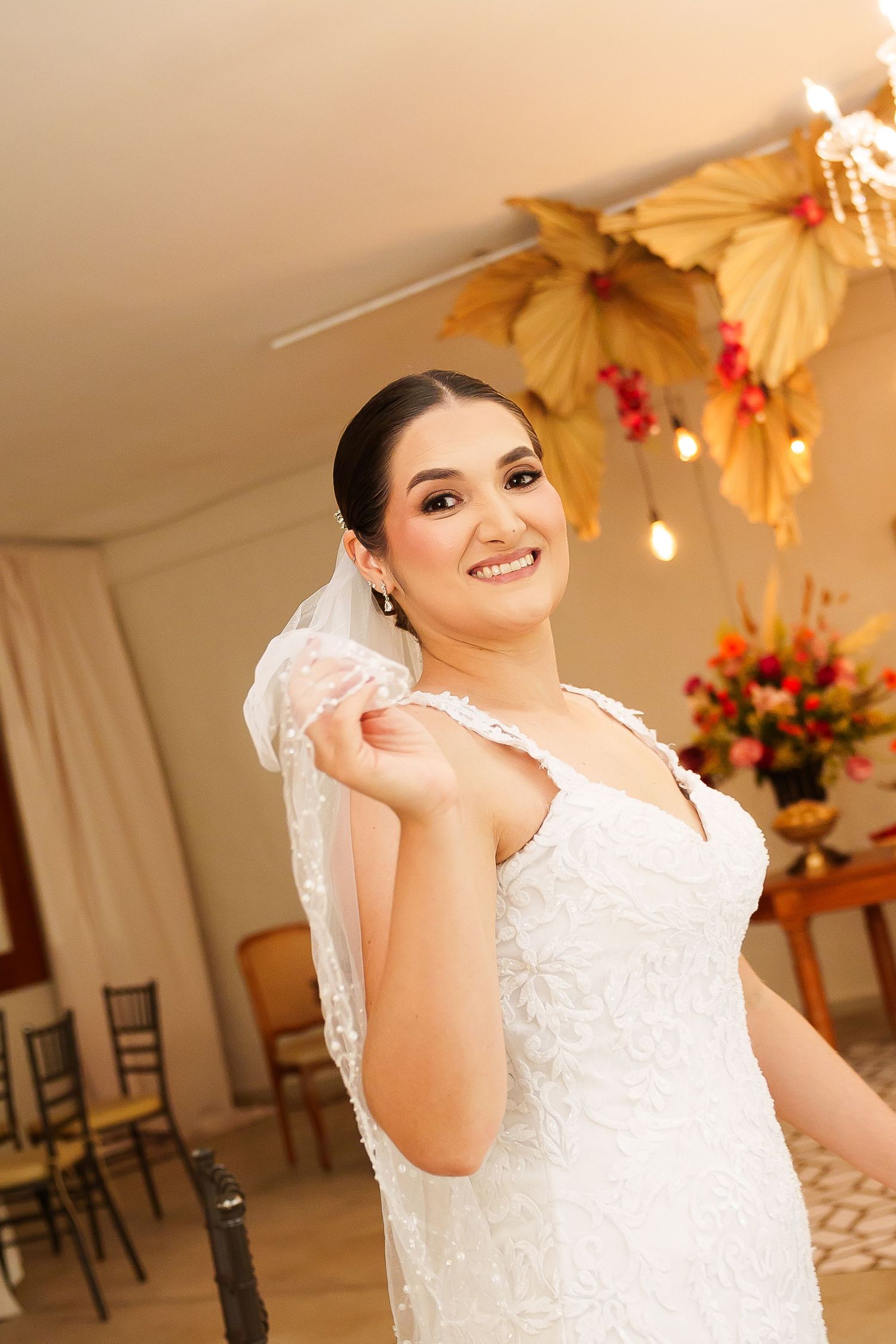 Smiling bride in white lace wedding dress holding veil, posing in elegantly decorated venue with floral arrangements.