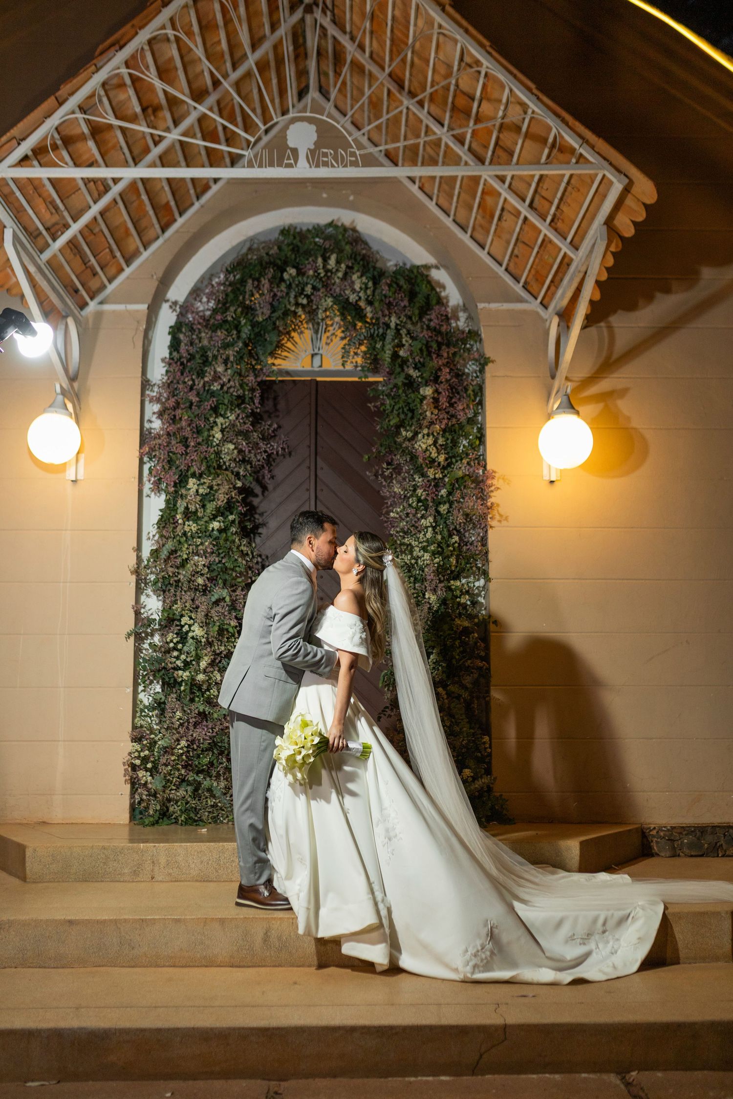 Bride and groom share a romantic kiss on church steps at night, framed by a floral arch and warm lights.