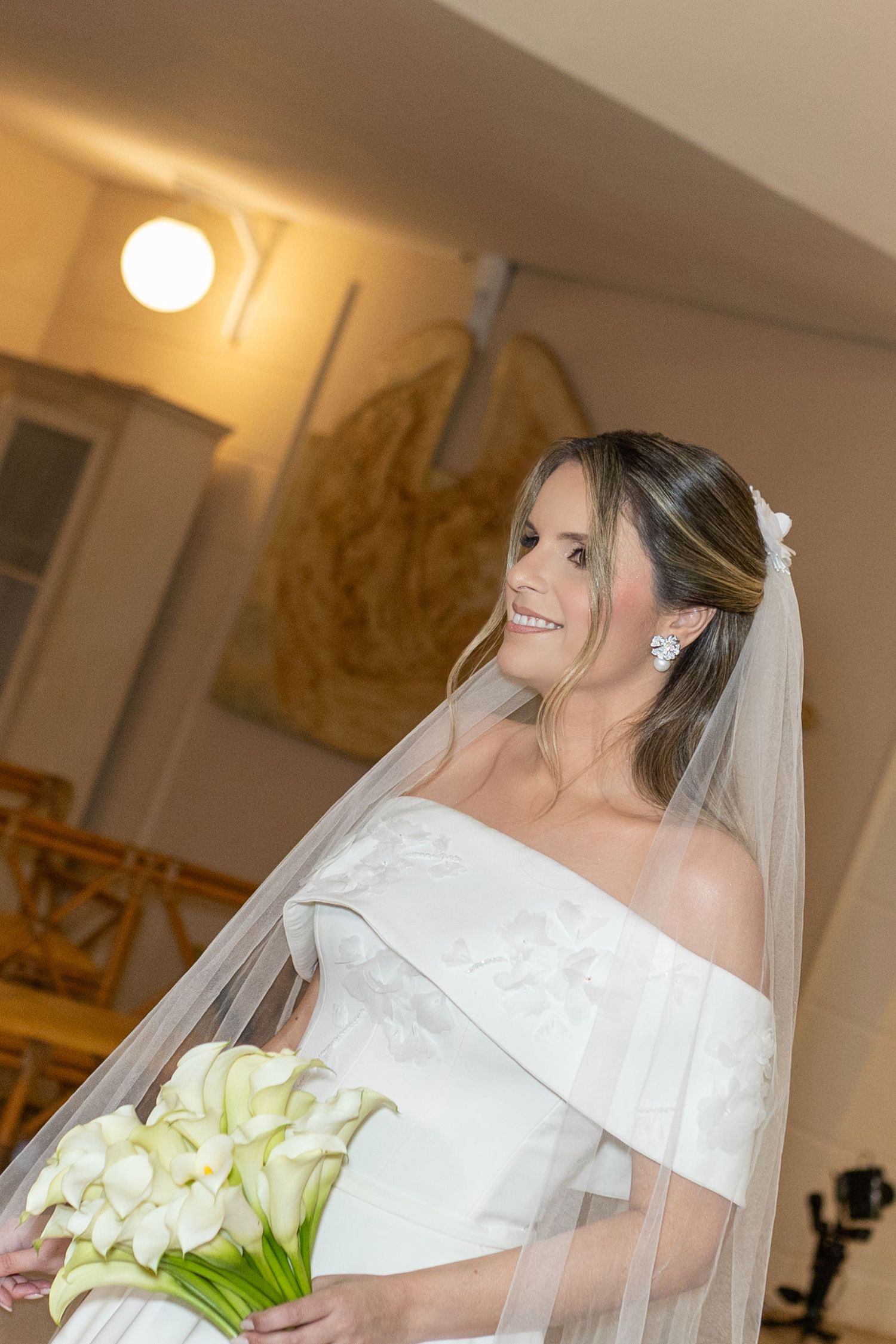 Smiling bride in off-shoulder white gown holding calla lily bouquet with veil and floral earrings indoors.