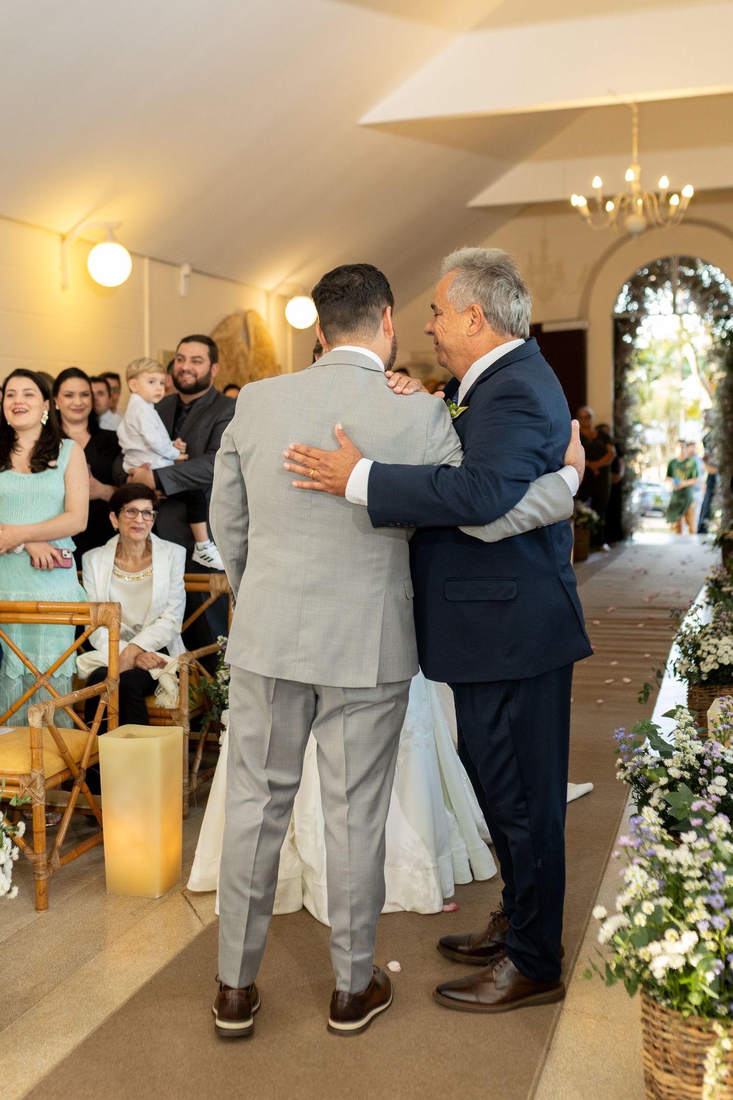 Groom embracing an older man in a suit during an indoor wedding ceremony with seated guests in background.