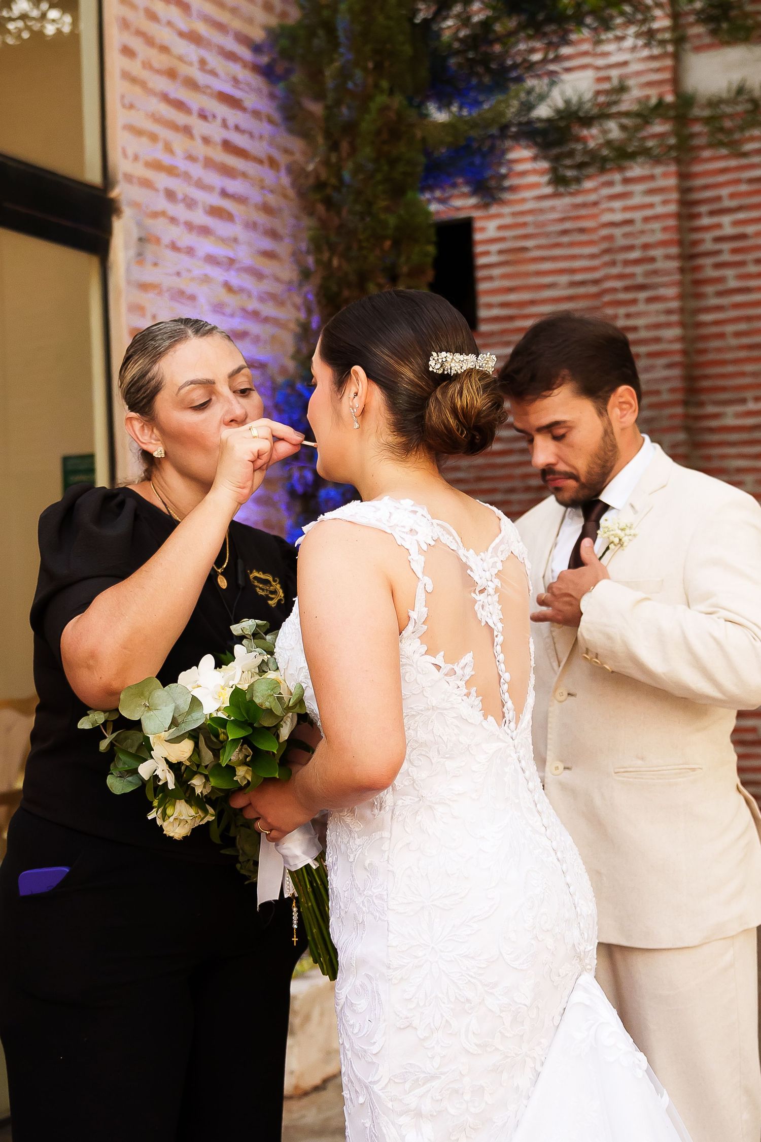 Bride in lace wedding gown holds bouquet while bridesmaid and groom adjust details before ceremony.