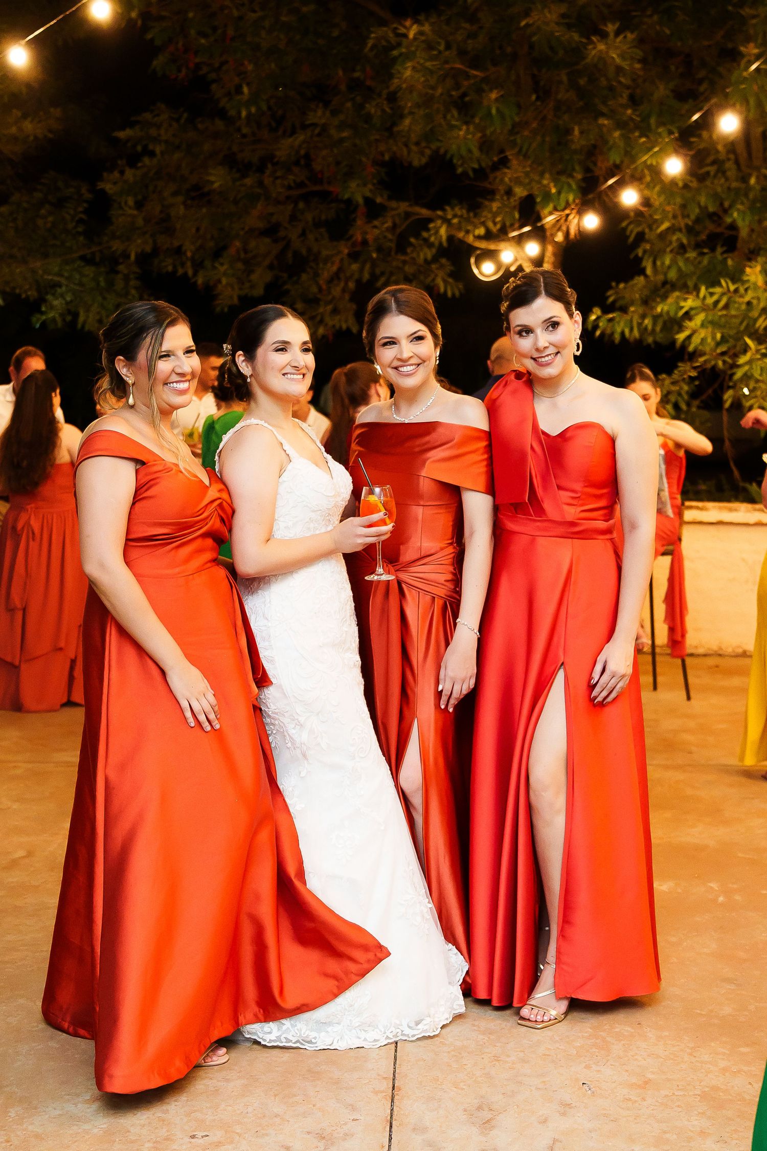 Bride in white lace gown poses with three bridesmaids in orange-red satin dresses at outdoor evening wedding reception.