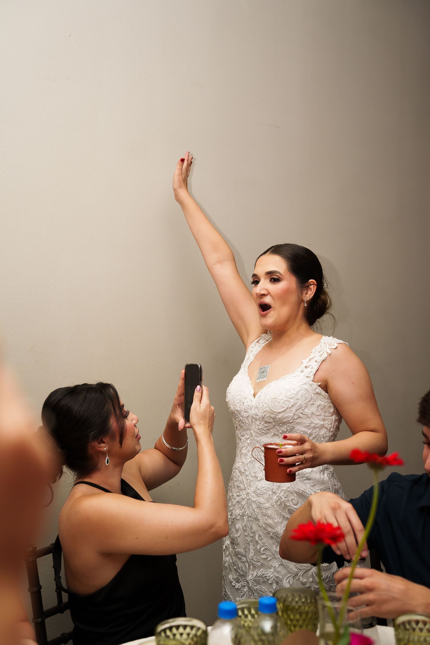 Bride in white lace gown raises arm celebrating at wedding reception while guest photographs her with smartphone.