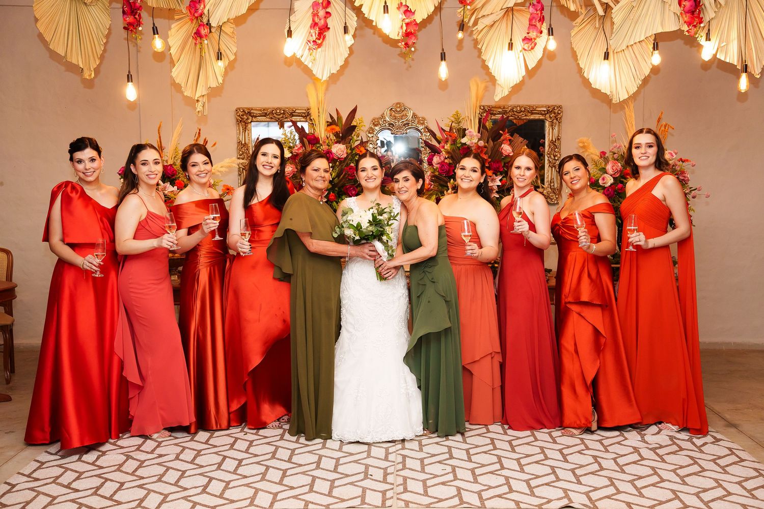 Bride in white gown poses with bridesmaids in red and green dresses at elegant floral wedding reception.