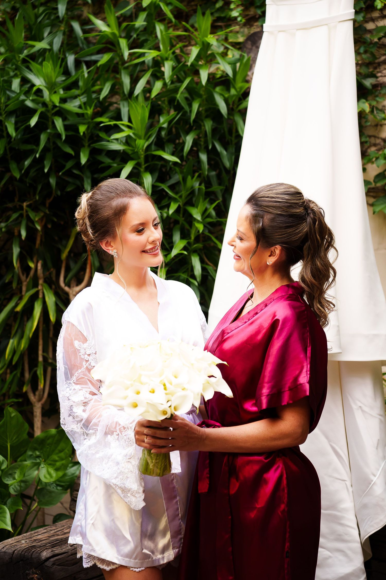 Bride in white robe holds bouquet with bridesmaid in red robe, sharing a joyful moment outdoors.
